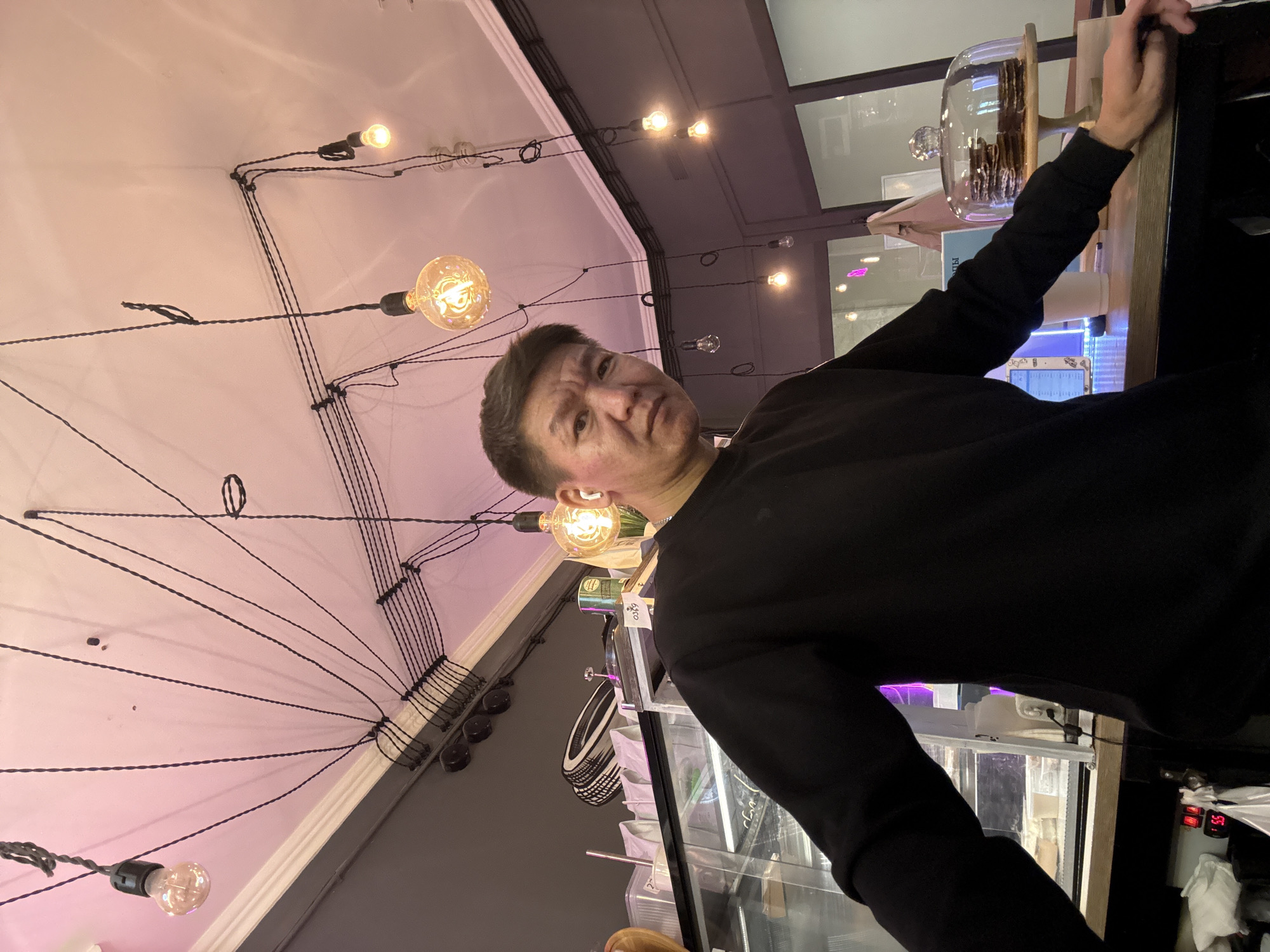 A man is taking a selfie in what appears to be a cafe. He is wearing a black sweatshirt and white earbuds. The background shows a ceiling with exposed wires and multiple Edison-style light bulbs hanging down. There are also glimpses of a display case with food items and a counter with a cake dome. The lighting is warm and ambient.