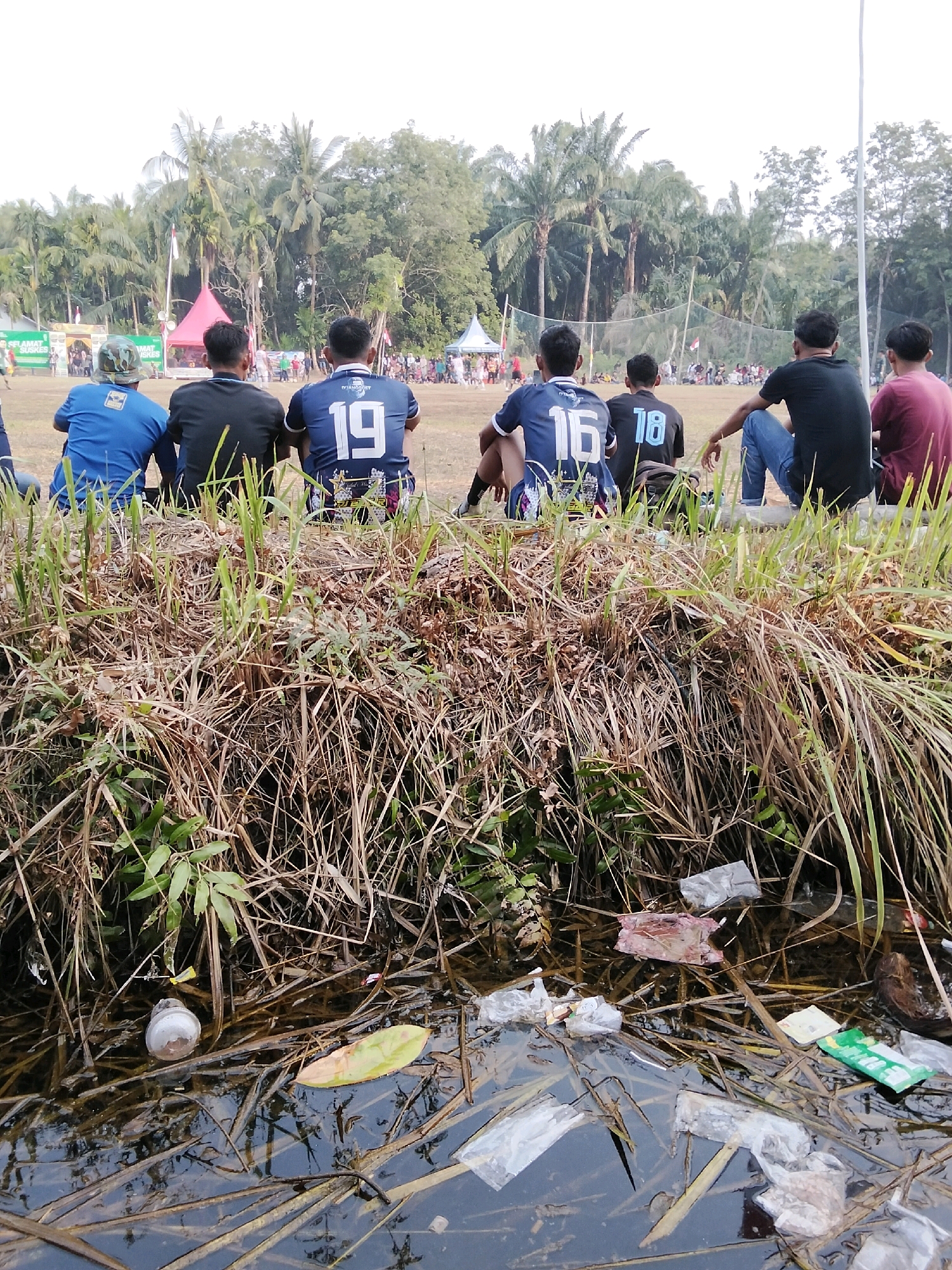 In Pambang Pesisir, Indonesia, a group of young men, likely a sports team, are sitting on a grassy bank, facing away from the camera. They are wearing athletic jerseys with numbers: 19, 16, and 18 are clearly visible. The foreground is dominated by dry grass and vegetation, with a small, murky body of water reflecting the scene. Scattered in the water and along the bank are various pieces of trash, including plastic bottles, wrappers, and other debris, indicating a lack of environmental cleanliness. In the background, a soccer field is visible with a crowd of spectators and tents, suggesting a sporting event is in progress. Palm trees and other lush vegetation form a dense backdrop, characteristic of the tropical Indonesian climate. The overall impression is of a casual outdoor gathering during a sporting event, marred by the presence of litter.