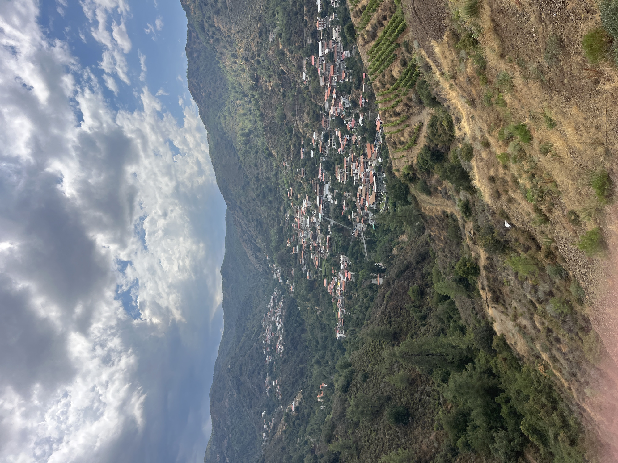 This is an aerial, high-angle, medium shot of the village of Kalopanagiotis, Cyprus, nestled within a lush, green, mountainous valley. The village itself is densely populated with buildings featuring terracotta-colored roofs and white walls, spread across the slopes. A winding road can be seen entering the village from the right side of the frame. In the foreground, a steep, dry, grassy slope dominates the lower half of the image, transitioning into the denser vegetation of the valley. Further up the slopes, terraced vineyards are visible, with neat rows of green vines. The sky above is partially cloudy, with dramatic grey and white clouds interspersed with patches of blue sky. The overall impression is one of a picturesque, rural village situated in a dramatic natural landscape. There are no people visible in the image.
