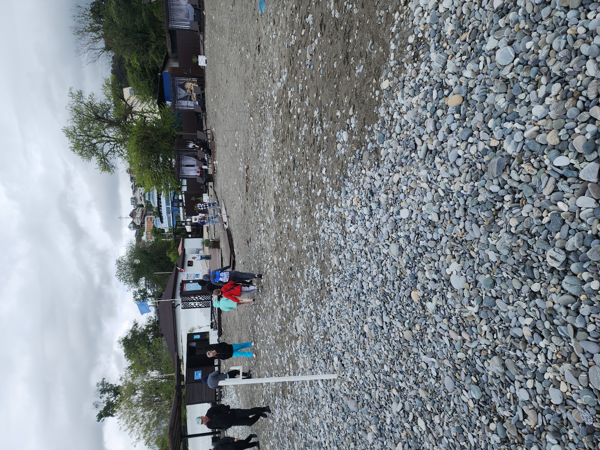The image shows a view of a beach with mostly grey pebbles, in Sochi, Russia. Buildings and trees run along the left side of the frame. Several people are standing or walking along the beach near the buildings. A blue flag is visible in front of one building. The sky is overcast.