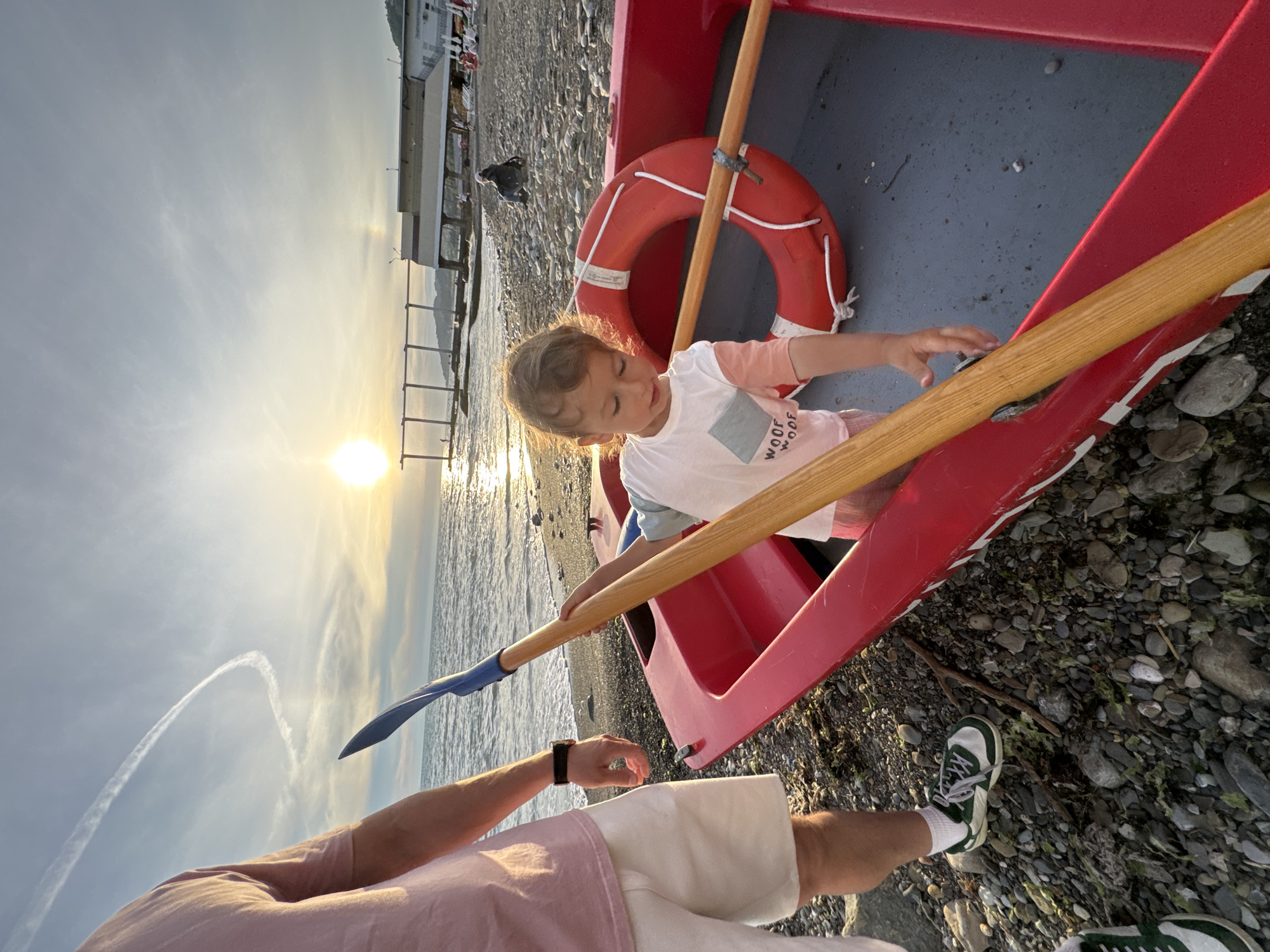 A young child is inside a red rowboat on a pebbly beach in Sochi, Russia, holding a wooden oar with a blue paddle. The child is wearing a white t-shirt with a graphic design. A person's legs, wearing white shorts and green and white sneakers, are visible on the left side of the frame. A red life preserver is inside the boat. The sun is low in the sky, casting a warm glow on the scene, and reflecting on the water. The sea is visible in the background, with a pier and some buildings along the coastline. The sky has contrails from an airplane.