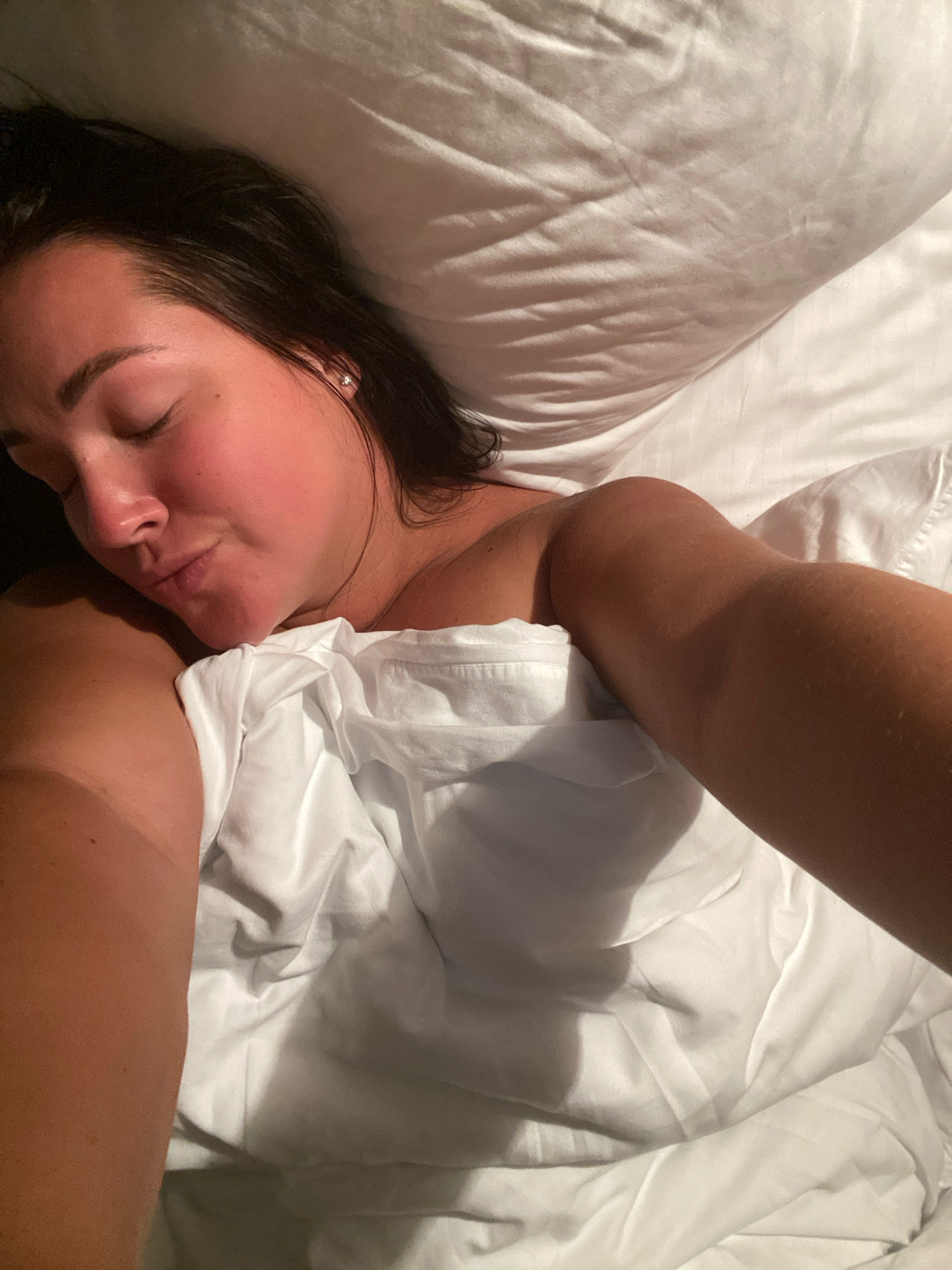 A woman is lying in bed with her eyes closed. Her head is tilted slightly to the side, and her lips are parted. She is wearing a white top that is partially covered by a white sheet. Her right arm is visible, and it appears to be resting on the sheet. The background is a white pillow. The scene is dimly lit, with shadows cast on the pillow and the woman's face. The image appears to have been taken in the city of Nebug, Russia.
