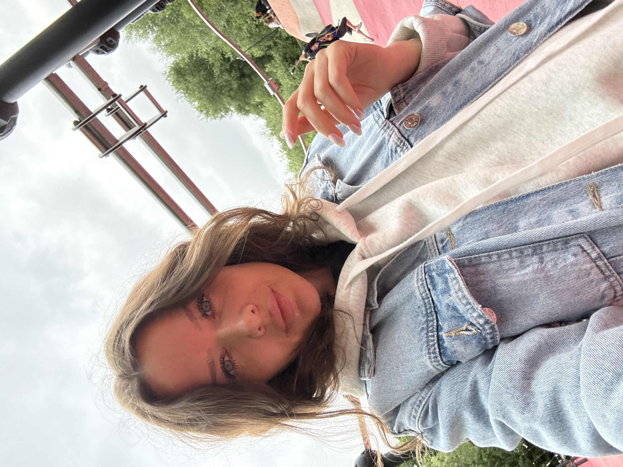 The image shows a young woman taking a selfie with a slightly downward angle. She has light brown hair, blue eyes, and is wearing a light blue denim jacket over a white hooded sweatshirt. Her right hand is visible, with manicured nails, resting near her chin. The background suggests an outdoor recreational area with a bright pink surface, trees, and metal structures that appear to be part of a playground or amusement ride. In the distance, other people are visible, some interacting with an elevated ride. The sky is overcast. The location is not specified as Moscow, Russia.