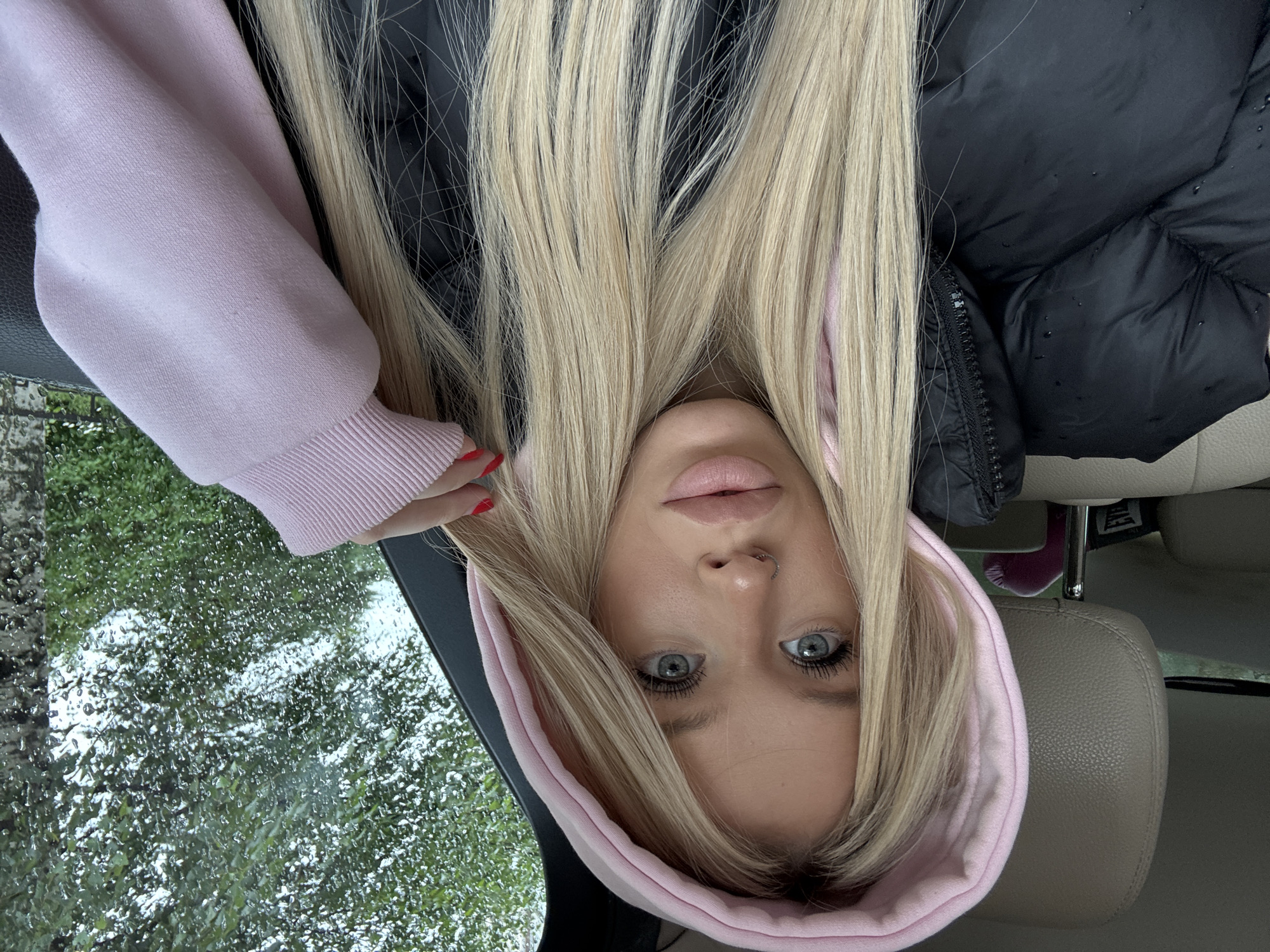 The media file is a selfie of a young woman taken inside a car. She has long, blonde hair, blue eyes, and is wearing a pink hooded sweatshirt. She has a nose ring and red nail polish. The image is taken from a low angle, with the woman's face appearing upside down. Outside the car window, it appears to be raining, with water droplets on the glass and greenery visible. The interior of the car is visible, including a leather seat and a portion of a black jacket. The woman's expression is neutral. There is no visible text in the image. The location context of Saint Petersburg, Russia, suggests the weather may be typical for the region.