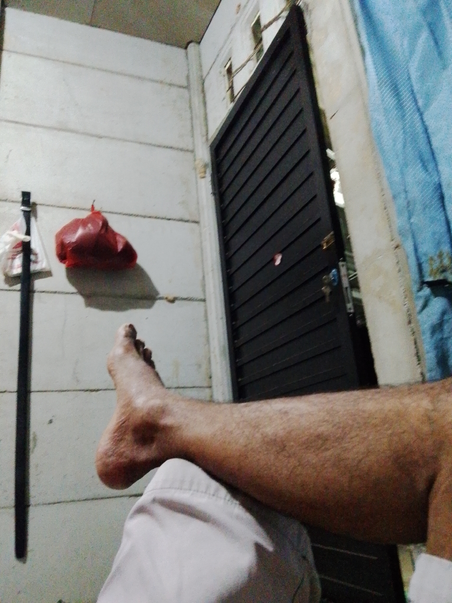 The image shows a portion of a person's leg, resting on what appears to be light-colored clothing, indoors. The background includes a white wall, a black, slatted door slightly ajar, and a blue curtain. A black belt hangs on the wall, along with a red plastic bag and a white plastic bag. The location is Bukit Ketok, Indonesia.