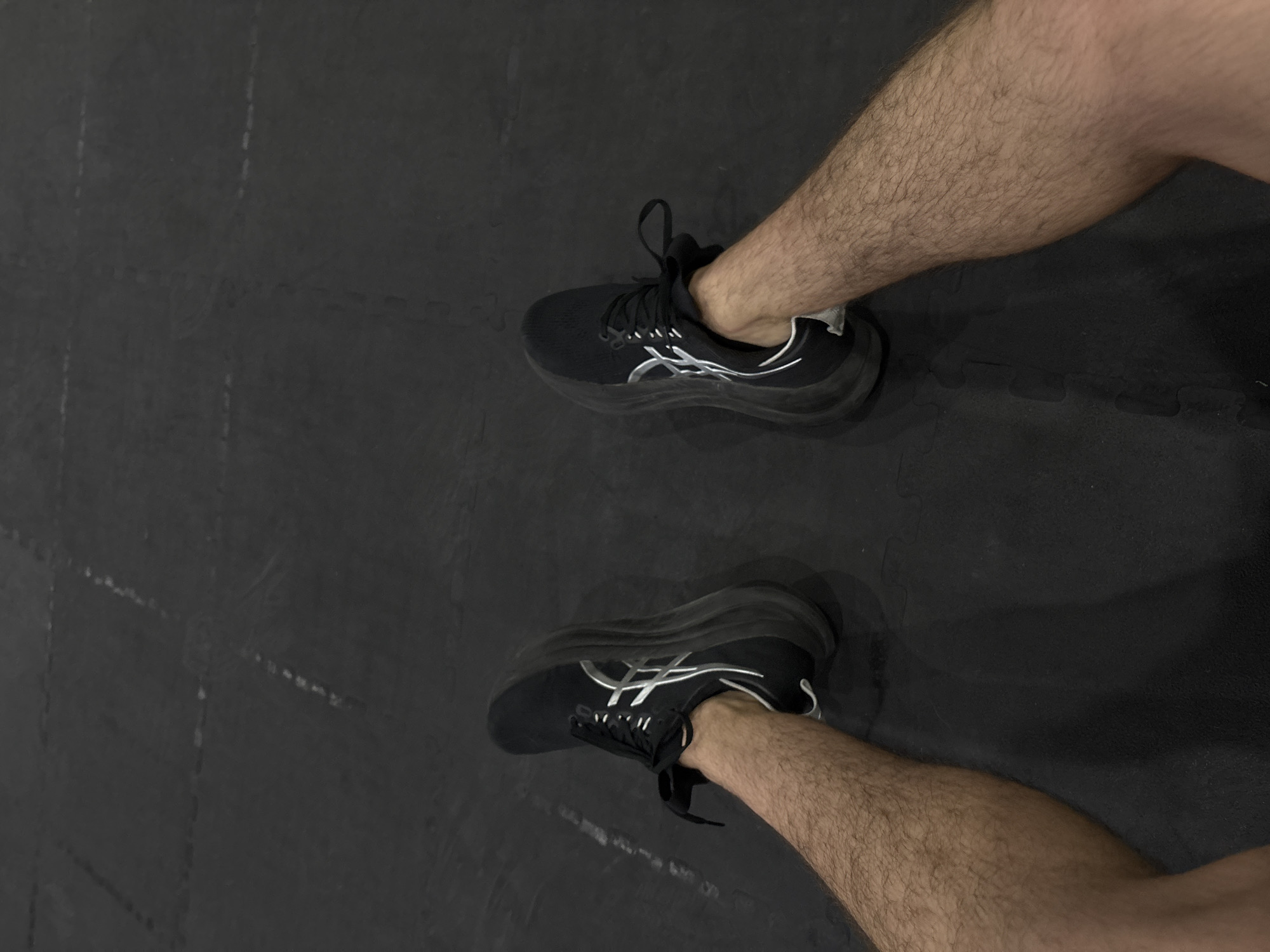 A downward shot shows a person's legs and feet clad in black running shoes with white accents, positioned on a dark grey interlocking tile floor. The legs are light-skinned and covered in dark hair. The scene is indoors. No additional information about San Bernardino Tlaxcalancingo, Mexico is available from the image.