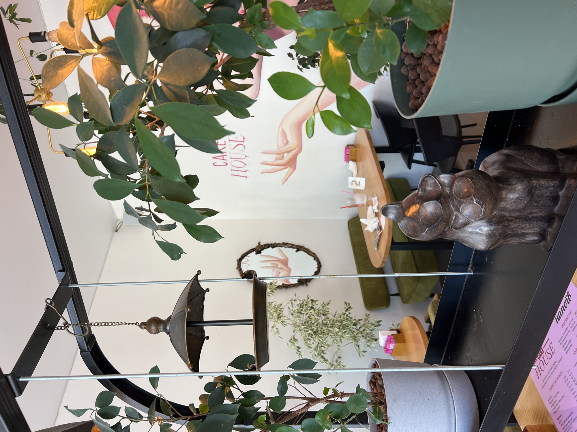 The image shows the interior of a cafe called "Cake House" in Odesa, Ukraine. In the foreground, a dark brown, cat-shaped sculpture sits on a black shelf. To the left, a tall plant in a light grey pot is visible. Behind the sculpture and plant, a mirror reflects a part of the cafe, including a table with drinks and a small sign with the number "2". The wall behind the table features a mural of a pink hand reaching out, with the words "CAKE HOUSE" written in pink lettering above it. Two green benches and a wooden table are visible in the mid-ground, along with another plant with variegated leaves. A metal bird feeder hangs from the ceiling in the left mid-ground. In the background, more plants and cafe seating are implied. The lighting suggests it is daytime.
