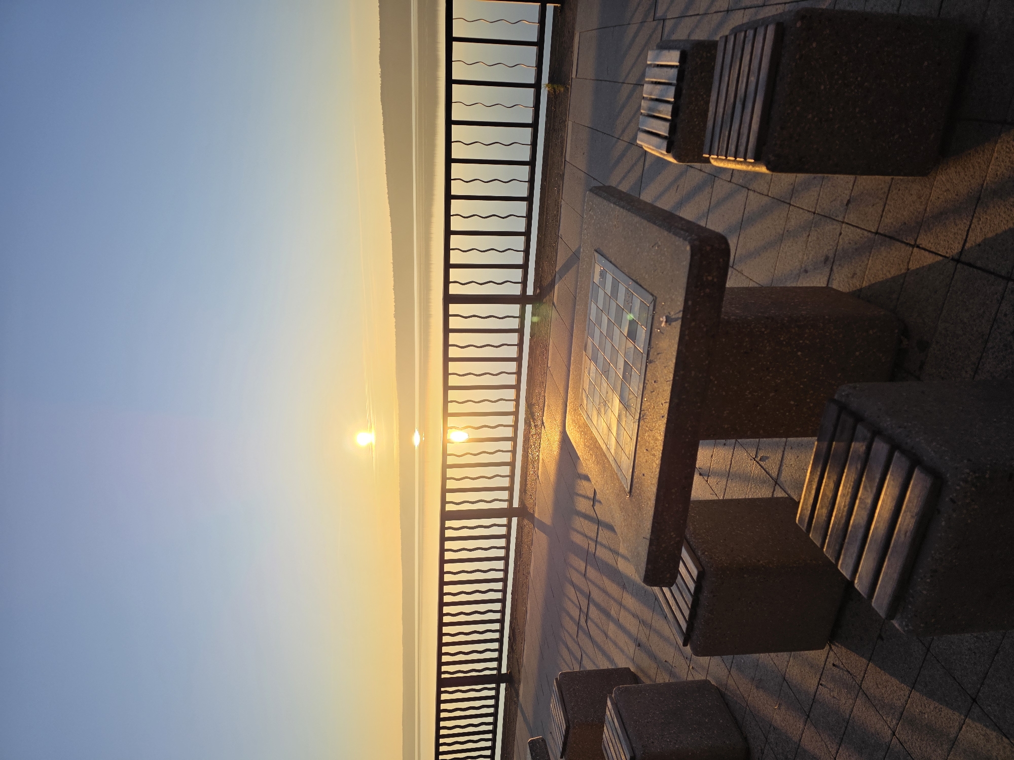 This image captures a serene sunrise or sunset scene along a waterfront promenade in Chaikovsky, Russia. The low sun casts a warm, golden light across the entire scene, creating long, dramatic shadows.
In the foreground, on the right side, there is a stone table with an inlaid chessboard or checkerboard, brightly illuminated by the sun. Several sturdy stone benches with wooden slats are arranged around the table and along the promenade. These objects cast prominent, elongated shadows across the grey paved ground, which consists of square tiles.
A dark metal railing with distinctive wavy horizontal bars runs horizontally across the midground, separating the paved area from a vast body of water. The railing itself casts shadows onto the promenade. Beyond the railing, the sun is visible as a brilliant golden orb, positioned low on the horizon, with its reflection stretching across the calm surface of the water.
In the far distance, across the expansive water, a faint, dark line indicates a distant landmass or shoreline, possibly hills or mountains. The sky transitions smoothly from a soft blue at the top of the frame to a warm, golden-orange hue near the horizon, indicative of either early morning or late evening.
There are no people visible in the image, contributing to a sense of tranquility and peacefulness. No visible text is present. The setting appears to be a public area designed for leisure and outdoor activities, like playing chess by the water.