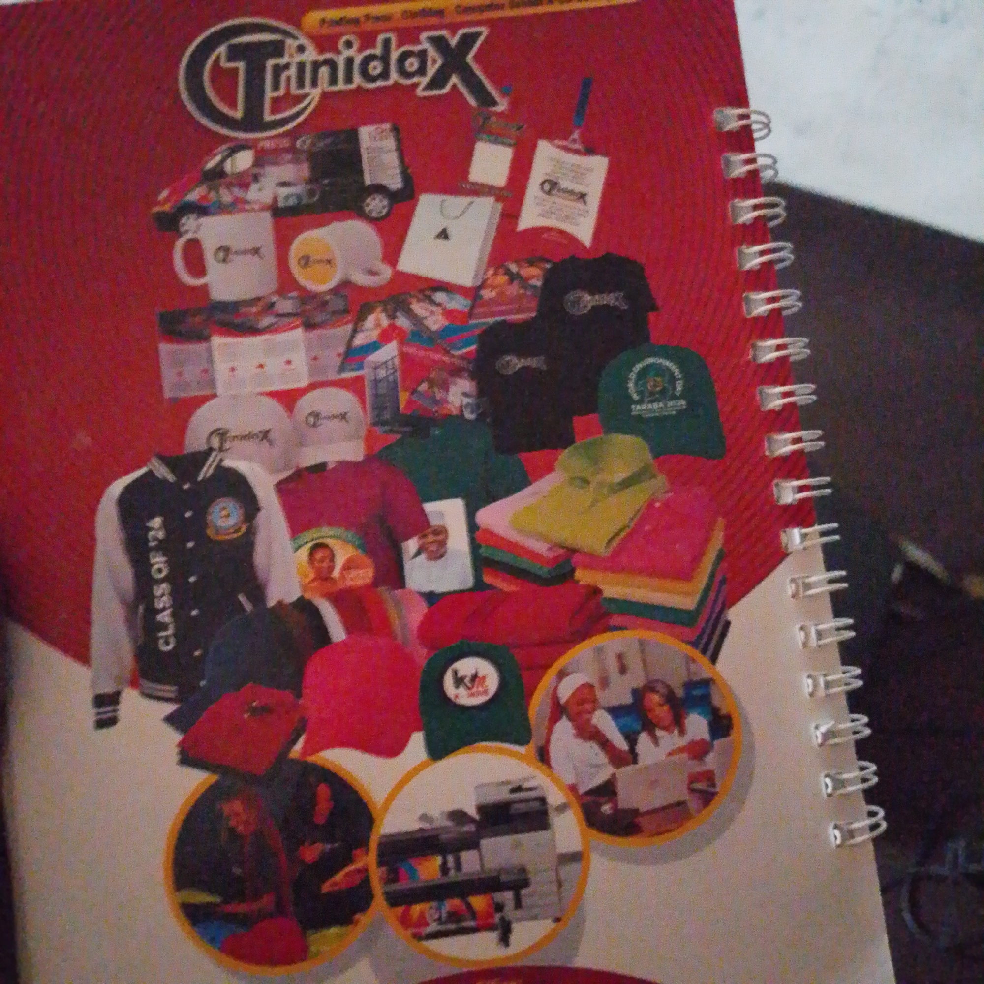 The image is a close-up, overhead shot of a catalog or brochure, likely for a printing and promotional items business, featuring a variety of merchandise. The cover is bright red with a large stylized "T" logo and the word "TrinidaX" in white. Scattered across the page are images of t-shirts, hats, mugs, a jacket with "CLASS OF '24" printed on it, a van, and various printed materials like flyers and bags. Several circular insets show people interacting with computers, a printer, and a woman wearing a headscarf. The spiral binding of the catalog is visible on the right side. The image quality is grainy. The catalog appears to be located in Jalingo, Nigeria, as indicated by the text overlay.
