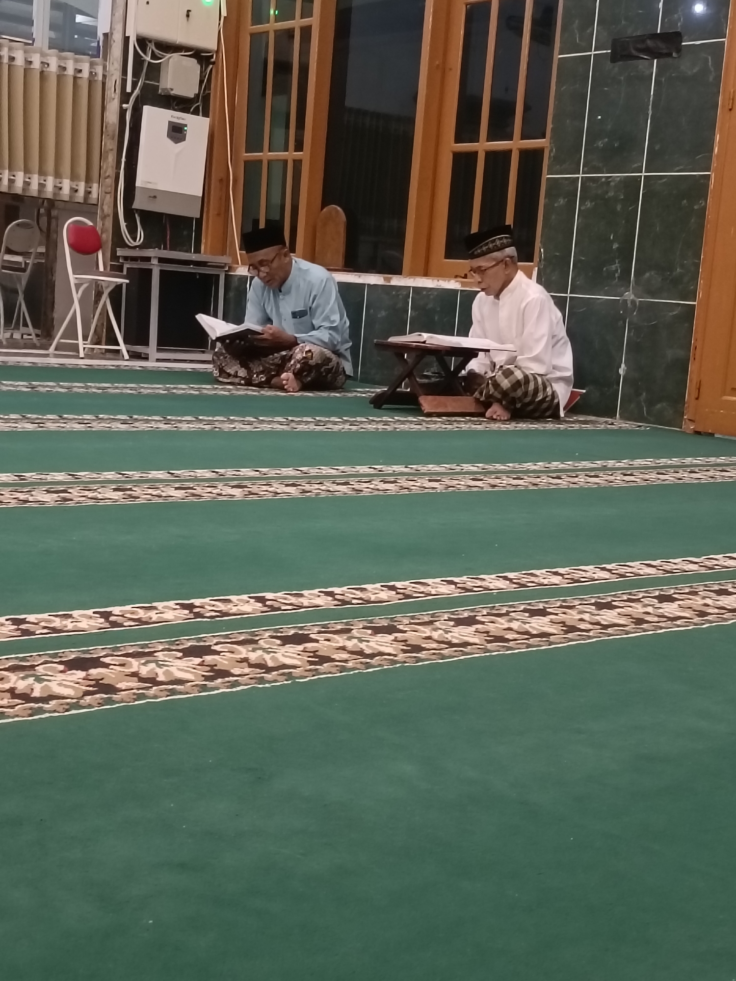 Two men are sitting on a green carpeted floor in a room with large wooden-framed windows and dark green tiled walls. Both men are wearing traditional Islamic attire, including kufi caps and patterned sarongs. The man on the left wears a light blue shirt, while the man on the right wears a white shirt. Both are reading from books placed on small wooden stands. The room appears to be a mosque or a prayer room, possibly located in Kediri City, Indonesia, given the context. The floor has decorative patterned bands. In the background, there are chairs, a table, and electrical equipment mounted on the wall.