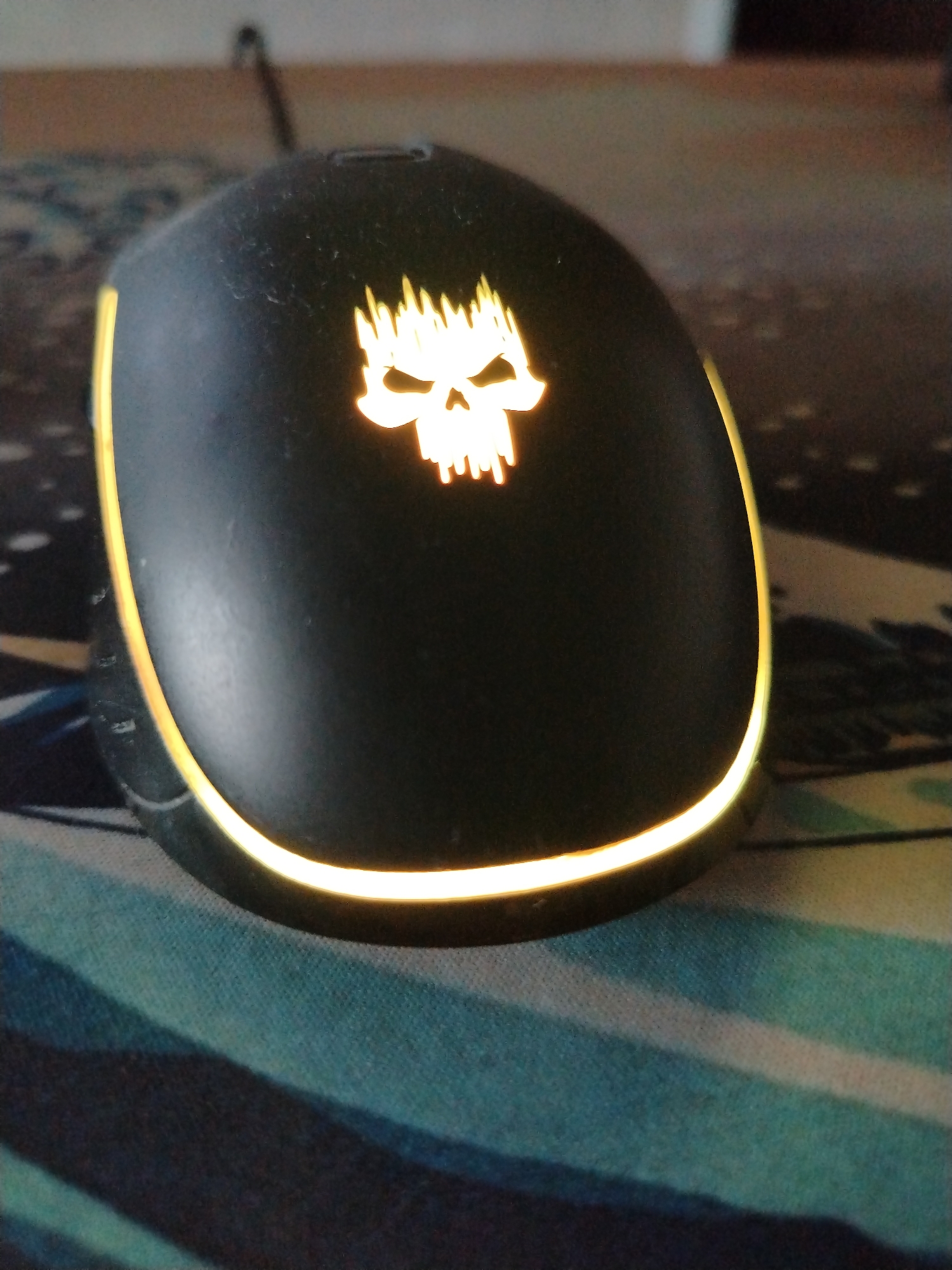 A black computer mouse with a glowing yellow skull emblem and a yellow illuminated ring around its base sits on a patterned mousepad. The mousepad has a mottled design with streaks of blue, teal, and black. The background is out of focus, showing a blurred surface and a hint of a cable extending from the mouse. The scene is indoors, likely on a desk.