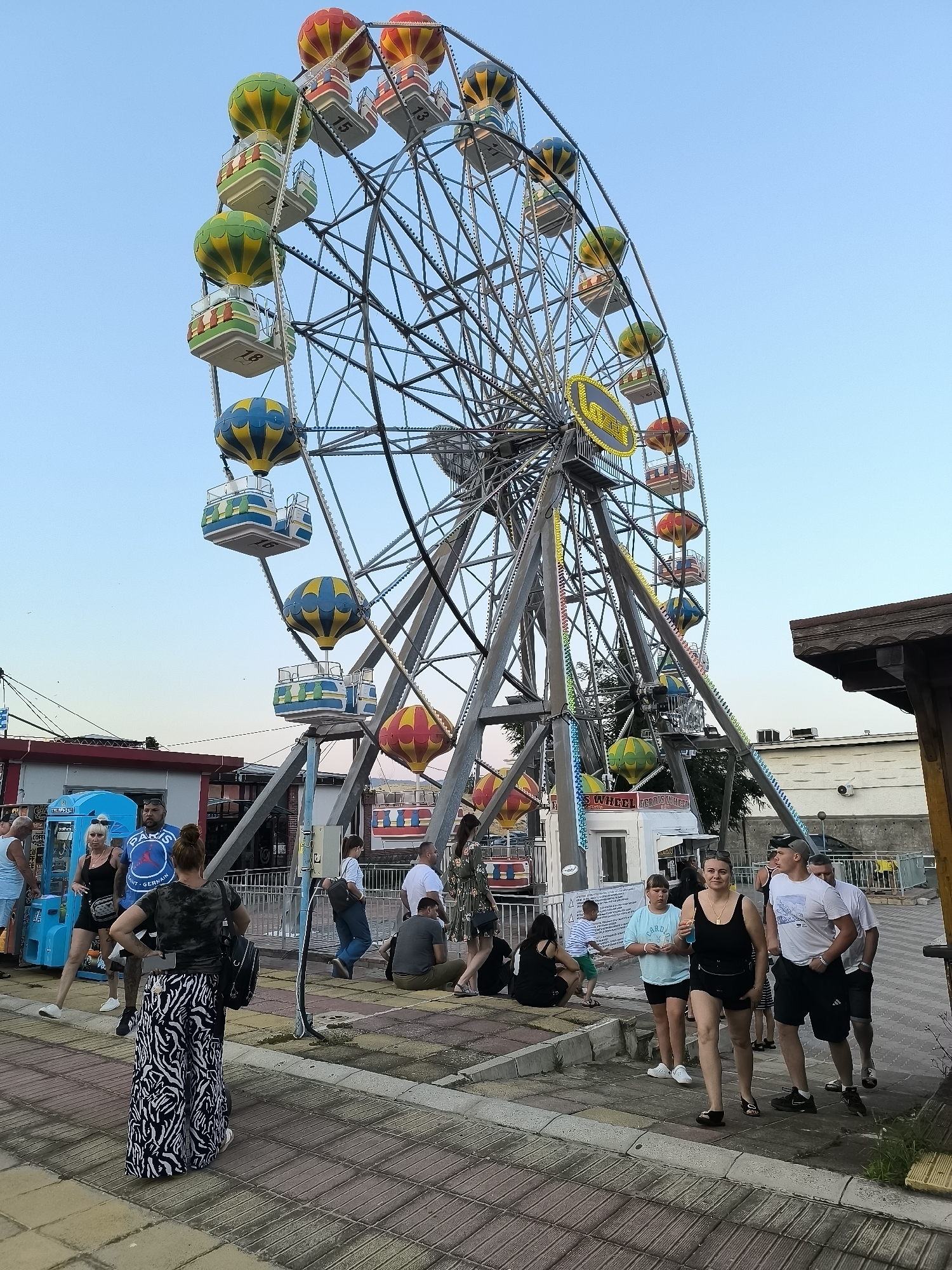 The image depicts a Ferris wheel with hot air balloon-shaped gondolas at an amusement park in Nesebar, Bulgaria. The Ferris wheel is the central and largest object in the frame, with its metal structure dominating the upper portion of the image. The gondolas are brightly colored in red, yellow, and green.
In the foreground and midground, several people are visible. On the left, a woman with dark hair tied back wears a camouflage t-shirt and a long, patterned skirt, and carries a backpack. She is looking to her right. Next to her, a woman in a black tank top and shorts and a man in a blue t-shirt with "Paris Saint-Germain" on it are standing.
Further to the right, a group of people are walking and standing. A woman in a black tank top and shorts is walking towards the camera, holding a drink. Beside her, a man in a white t-shirt and black shorts walks away from the camera. Other individuals are scattered throughout the scene, some sitting on a low wall.
The environment appears to be an outdoor public space, possibly a promenade or boardwalk, with a paved ground surface made of interlocking stones in shades of yellow, brown, and gray. To the right, part of a wooden structure with a thatched roof is visible, likely part of a food stall or kiosk. Behind the Ferris wheel, buildings and what appear to be other amusement park attractions are present. The sky is a clear, pale blue, indicating daylight, likely late afternoon or early evening given the lighting. The overall atmosphere suggests a lively public gathering.