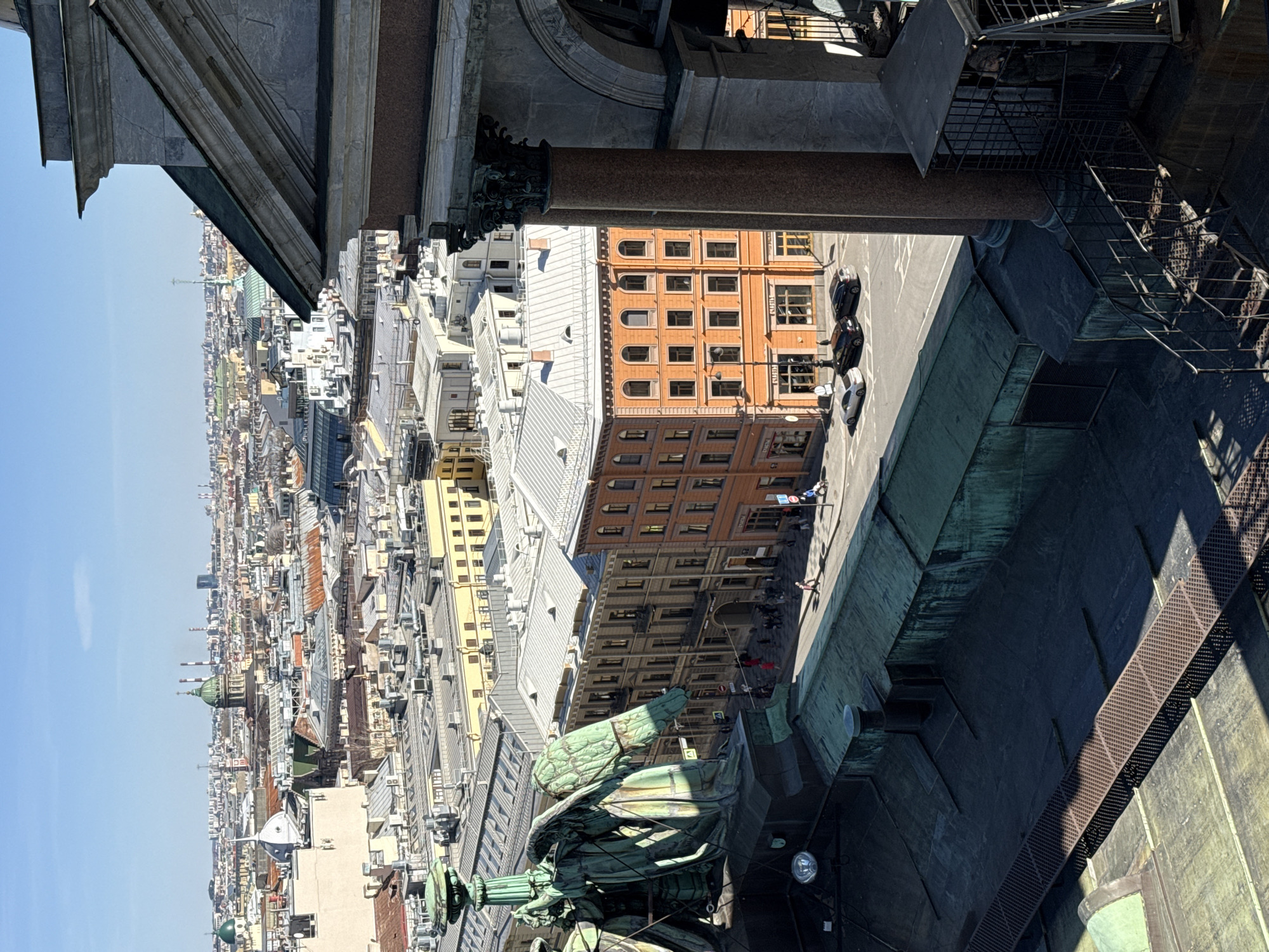 The image is an elevated view of Saint Petersburg, Russia, taken from the colonnade of St. Isaac's Cathedral. The architecture of the city is visible, including various buildings with different heights and colors. Some have ornate facades, while others are more plain. There's an orange building with many windows. A street is visible below with pedestrians and parked cars. The view from above offers a look at roofs, facades, and architectural details, as well as the street activity below. In the foreground are elements of St. Isaac's Cathedral, including statues and architectural supports. A metal staircase can be seen attached to the cathedral’s surface.