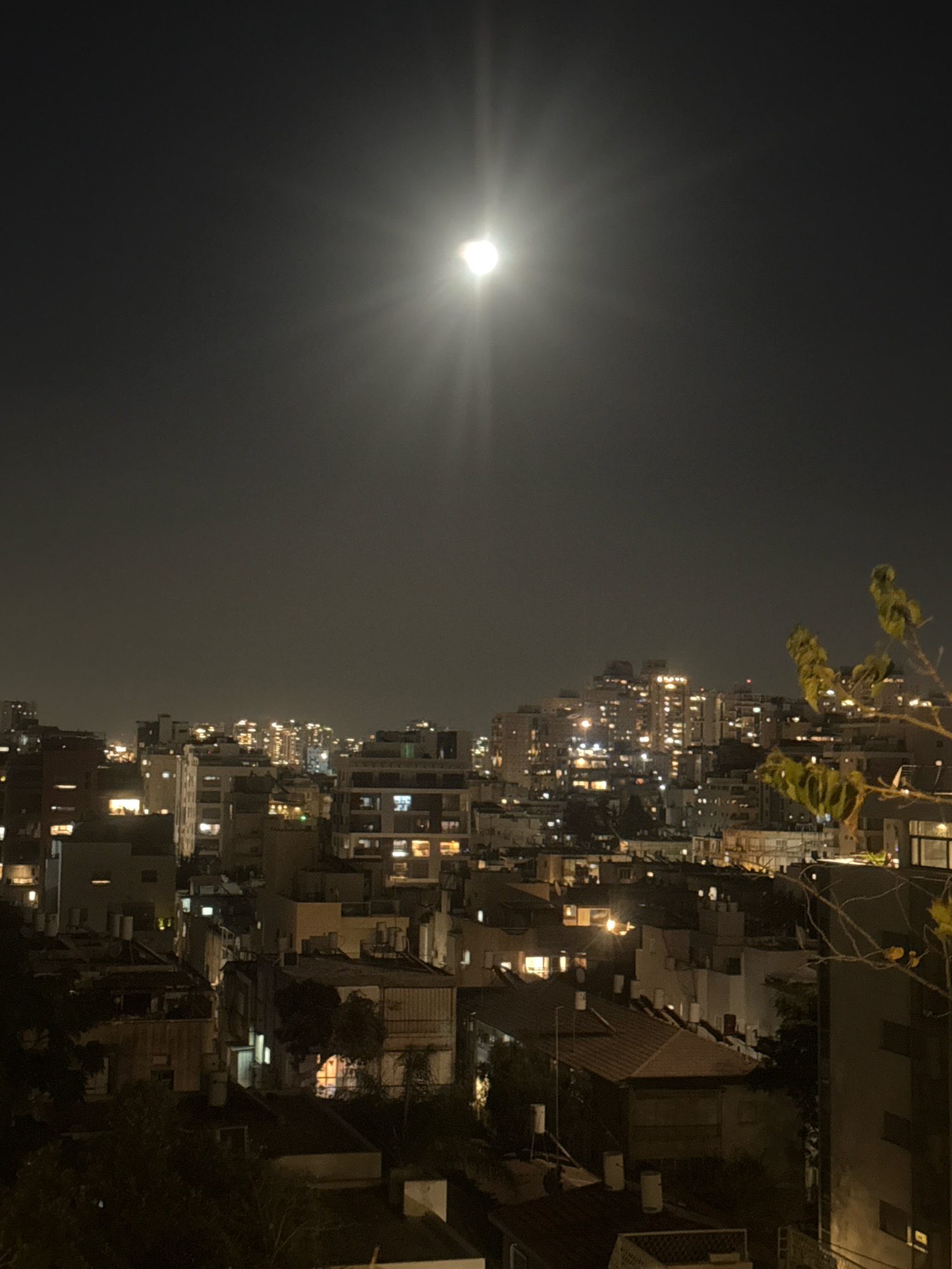 The image is a night-time photograph of a cityscape in Ramat Gan, Israel, under a full moon. The moon is prominently visible in the upper center of the frame, casting a bright glow and creating rays of light that extend across the dark sky. Below the moon, the city is illuminated by numerous lights from buildings, creating a warm, orange and yellow ambiance. The architecture appears to be a mix of residential and commercial buildings, with some appearing taller and more modern, while others are lower and more traditional. Trees and foliage are visible in the foreground and scattered throughout the city, adding a touch of nature to the urban landscape. The overall scene depicts a lively, inhabited city at night, bathed in the soft light of the moon. There are no people visible in the photograph.