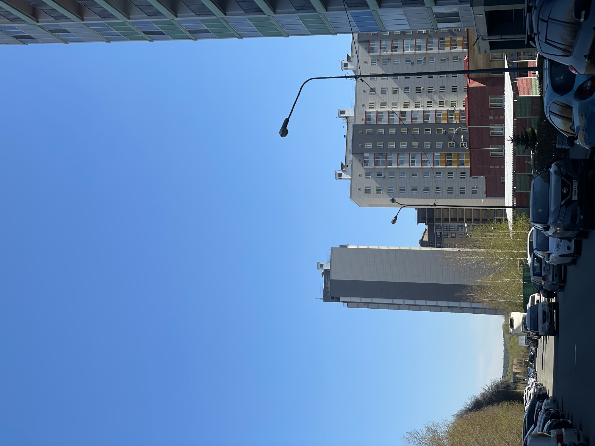 The image shows a street view in Kazan, Russia, lined with parked cars on the right. Tall residential buildings are visible on both sides of the street, with the sky above. Streetlights are interspersed along the road. One building has a mix of gray, yellow, and red colors on its facade, and another is predominantly gray. Bare trees and low-lying greenery are visible in the distance beyond the buildings. The sky is clear and blue.