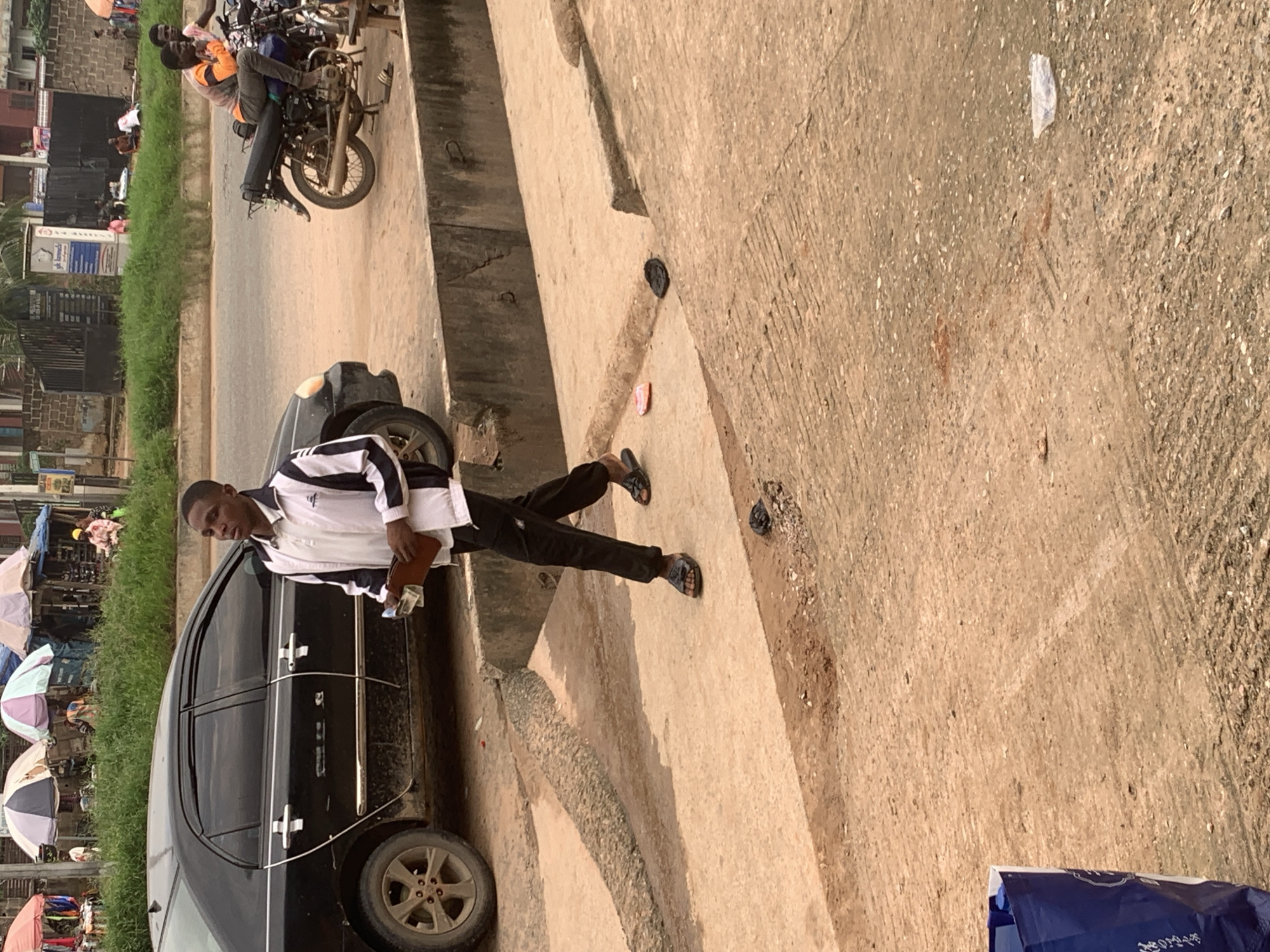 A young Black male wearing a white polo shirt with black and white striped sleeves and black pants walks on a concrete sidewalk. He is holding a brown wallet with cash sticking out of it in his right hand. In the background, a black sedan is parked on the street, and several people are visible under colorful umbrellas at a market. The scene appears to be in Shagamu, Nigeria, indicated by context clues such as the market scene, the type of vehicles, and the general environment. The ground is dry and dusty, with some scattered debris.