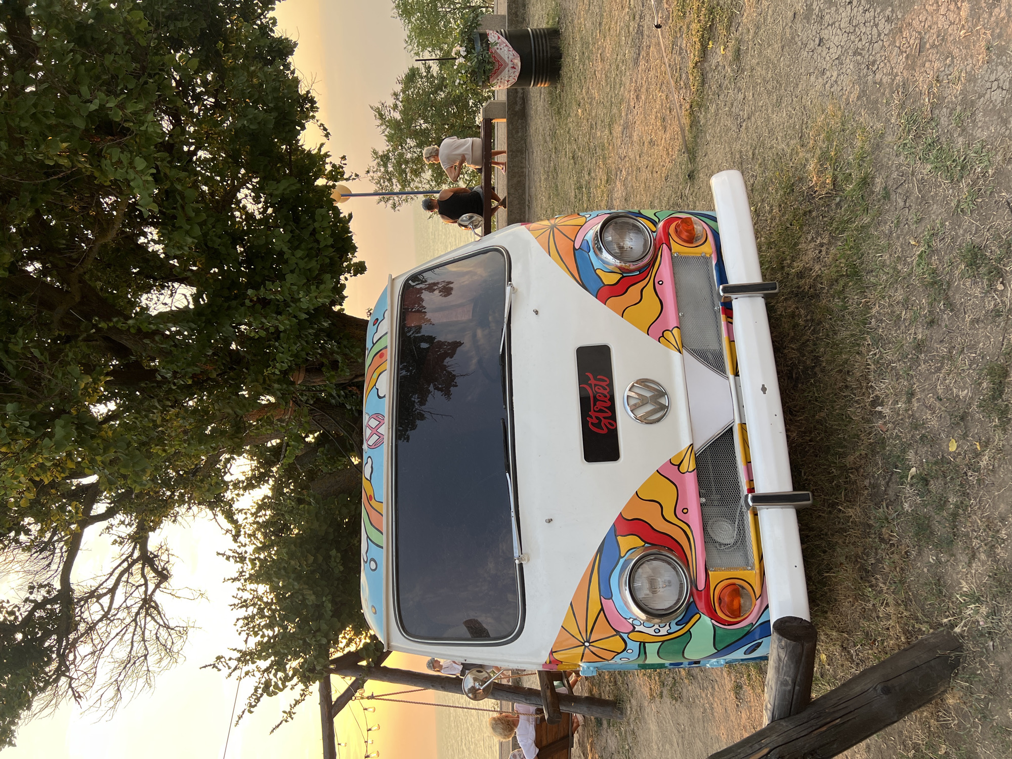 In Yeysk, Russia, a vintage Volkswagen van, vibrantly decorated with a psychedelic pattern of flowers, waves, and rainbow motifs, is parked on a grassy area near a body of water. The van features a white base with colorful decals adorning the front and hood. A license plate with the word "Street" in red script is affixed to the front.
The scene is set outdoors, likely during sunset given the warm, golden light illuminating the sky and casting long shadows. In the background, a large tree with lush green leaves provides shade. A wooden swing set is visible to the left, and further back, two people are seated on a bench, facing the water, suggesting a relaxed atmosphere. A black barrel planter with white flowers sits to the right. The ground is a mixture of dry grass and dirt. The overall impression is one of a casual, possibly festive, outdoor gathering by the sea.