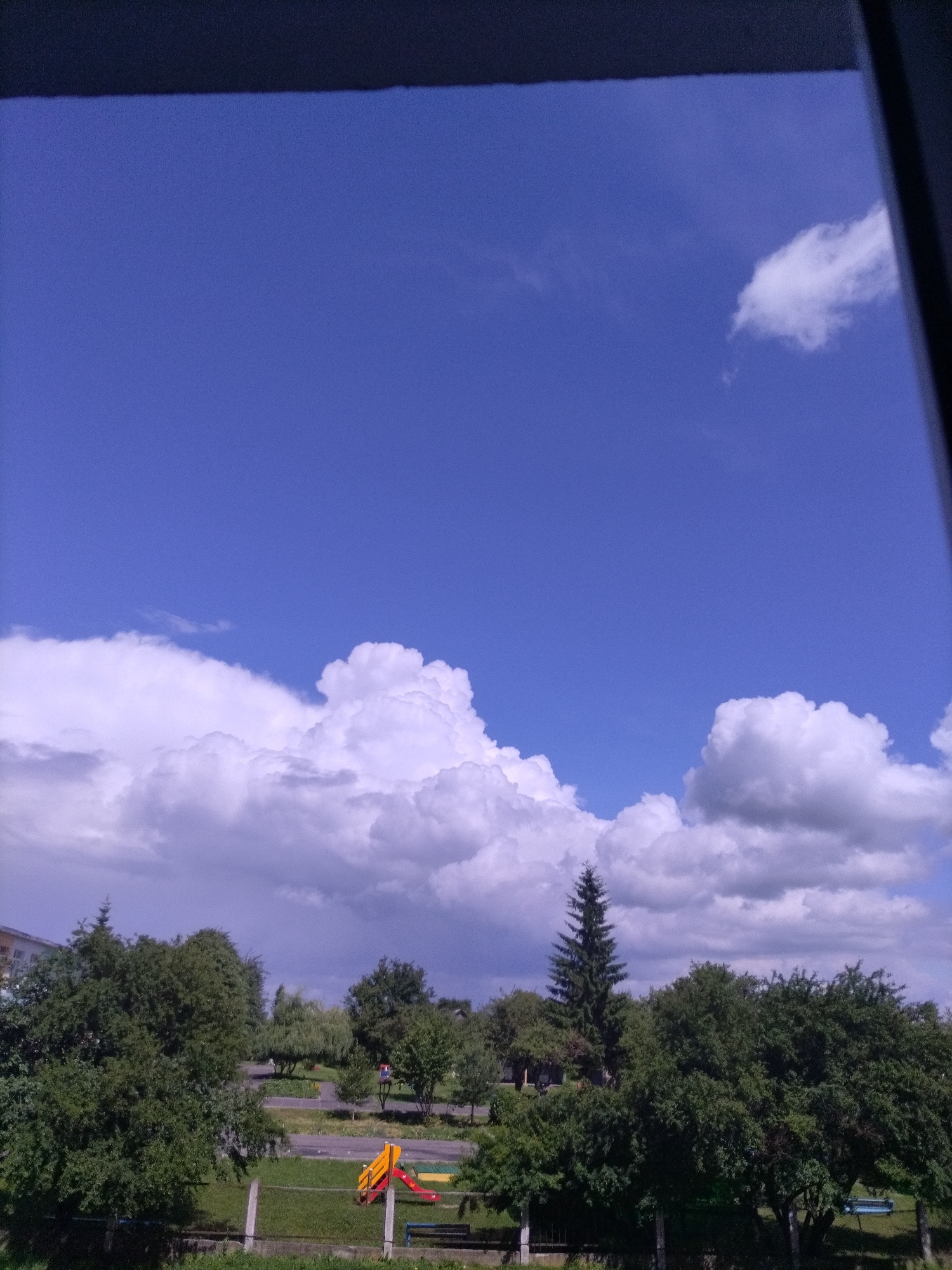 The image is a wide-angle shot taken from a slightly elevated position, looking out over a green landscape under a vast blue sky filled with fluffy white clouds. In the foreground, a dark structure, possibly a window frame or overhang, partially obscures the top of the image. Below this, the sky occupies the majority of the frame, transitioning from a deep blue at the top to lighter shades near the horizon. A single, small white cloud floats in the upper right quadrant.
The lower portion of the image reveals a park or recreational area. Dense green trees line the background, with a few interspersed buildings, one of which appears to be a multi-story apartment building on the far left. A paved pathway winds through the greenery, and a playground with a yellow and red slide is visible. A blue bench sits to the right of the slide, and a dark blue bench is visible further back. The ground is covered in grass, and a fence runs along the bottom of the frame, with several concrete posts. A tall, slender evergreen tree stands prominently in the middle-ground. The overall scene suggests a pleasant, possibly summery day in an urban or suburban park.