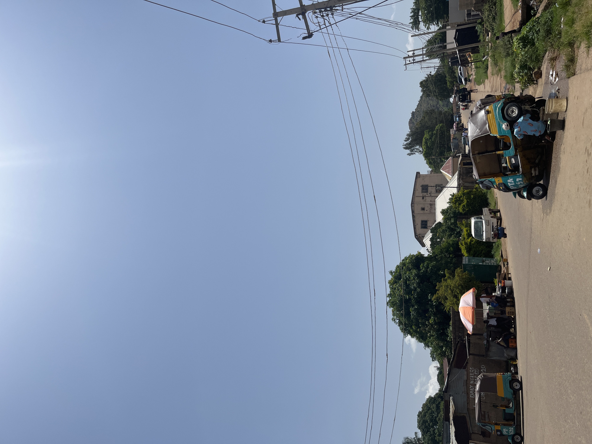 The image depicts a street scene in Gwong, Nigeria, on a clear, sunny day. The road is paved and appears to be a local thoroughfare with shops and residences lining one side.
In the foreground on the left, a turquoise and yellow auto-rickshaw, identified by the license plate "PTA 1692," is parked. Further down the road, another auto-rickshaw is the central focus. It is tilted on its side, and a person wearing a blue and white patterned shirt is seen washing it, with buckets and water present.
Along the left side of the road, a row of stalls or small shops with corrugated metal roofs are visible. An orange and white umbrella shades a vendor area where people are gathered. A white pickup truck is parked further down on the right side of the road.
Power lines crisscross the sky, supported by wooden utility poles, indicating the presence of electricity infrastructure. Vegetation, including trees and shrubs, is scattered throughout the scene, suggesting a semi-urban or rural setting within the city. Buildings of varying architectural styles, from simple structures to more substantial houses, are visible in the background. The sky is a bright, cloudless blue, and the sunlight casts shadows, indicating midday. The overall atmosphere is one of everyday life and activity on a Nigerian street.