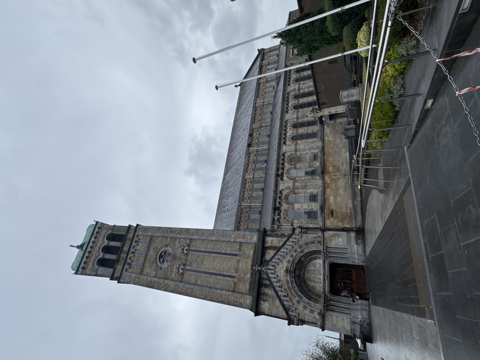 The image shows a wide-angle, low-angle view of St. Mary's Roman Catholic Church in Clonmel, Ireland, on an overcast day. The church is constructed of stone with a tall, square bell tower on the left, capped with a green copper roof. The tower has arched openings near the top and decorative stonework. The main body of the church features a long, sloping roof and multiple arched windows with stone surrounds.
In the foreground, a person is standing to the left of the church entrance, which is a large, ornate wooden door beneath an arched stone portico. The ground in front of the church is paved with dark stone tiles, appearing wet, suggesting recent rain. To the right, a metal ramp with handrails leads up to the church, bordered by a chain with red and white striped tape. Beyond the ramp, there is a garden area with neatly trimmed bushes and some gravestones visible, indicating a graveyard. Two tall, white flagpoles stand in the middle ground to the right, one slightly in front of the other. The overall atmosphere is somber due to the cloudy sky and wet conditions.