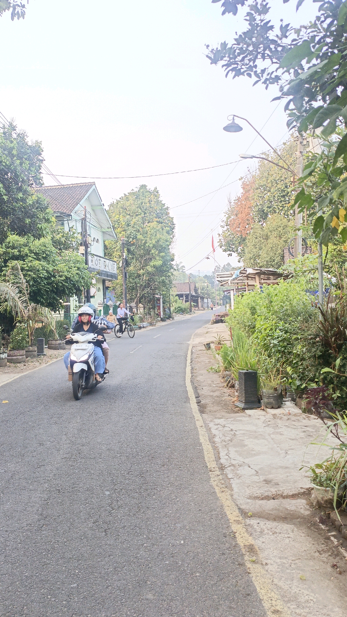 A motorcycle with two occupants travels on a paved road lined with vegetation and houses in Salatiga, Indonesia. The rider wears a helmet, and both individuals are dressed casually. A person on a bicycle is visible further down the road, also moving in the same direction. The environment suggests a rural or suburban setting with a mix of residential buildings and abundant greenery. The sky is overcast, and the lighting indicates it is daytime.