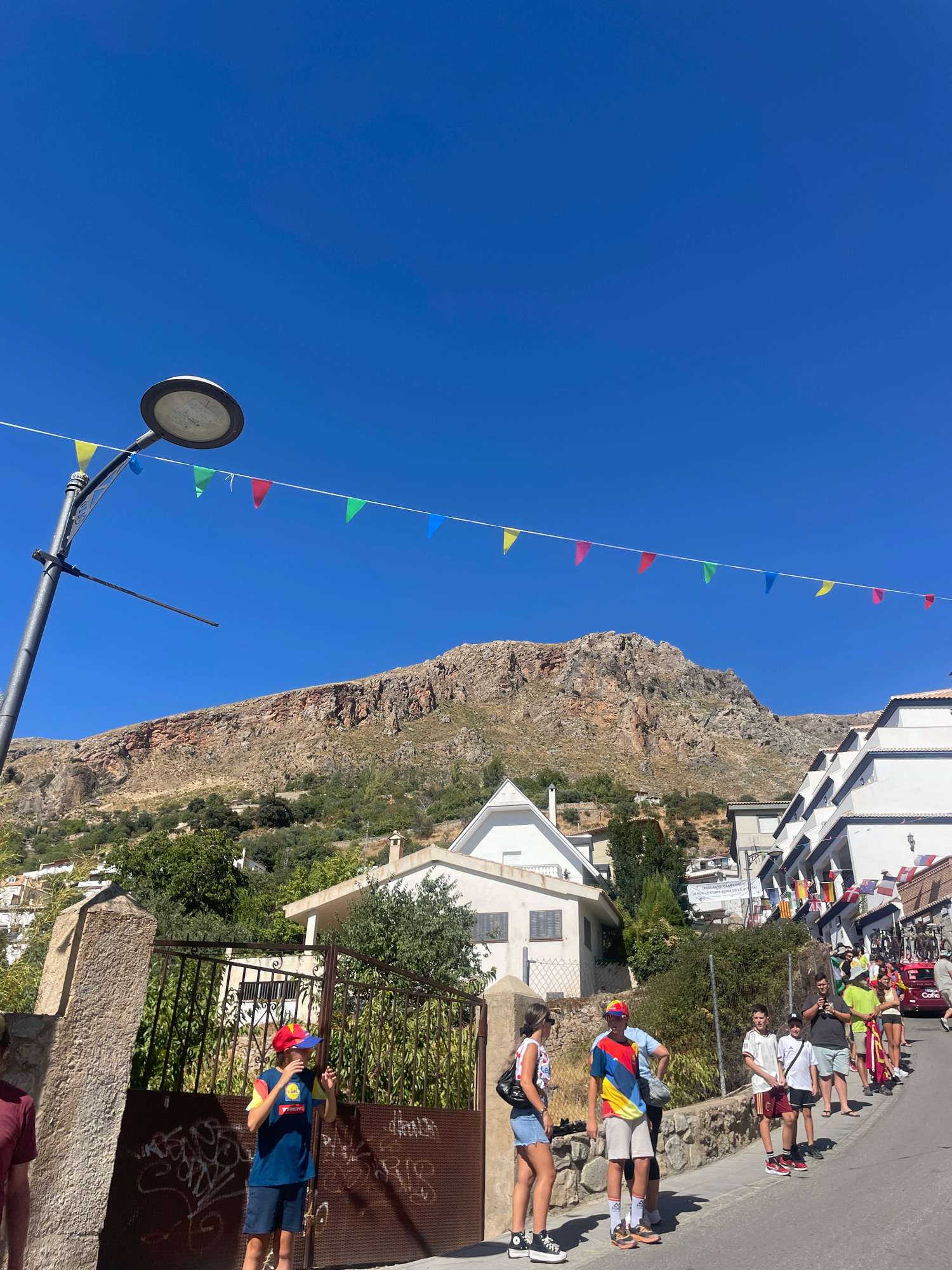 In Güéjar Sierra, Spain, on a clear, sunny day, a street is lined with buildings and a mountain looms in the background. A string of colorful triangular flags is strung across the sky, attached to a streetlamp on the left. Several people are standing on the right side of the street, some appear to be watching an event, possibly a race, as evidenced by bicycles on a nearby vehicle and flags draped across some buildings. A young boy wearing a blue t-shirt and a red and yellow hat is standing near a metal gate on the left side of the image, holding his hand up to his face. The buildings are white with red-tiled roofs, and some have balconies decorated with flags. The terrain is hilly, with trees and vegetation growing on the slopes. The street itself is paved, and there is a stone wall along the left side.