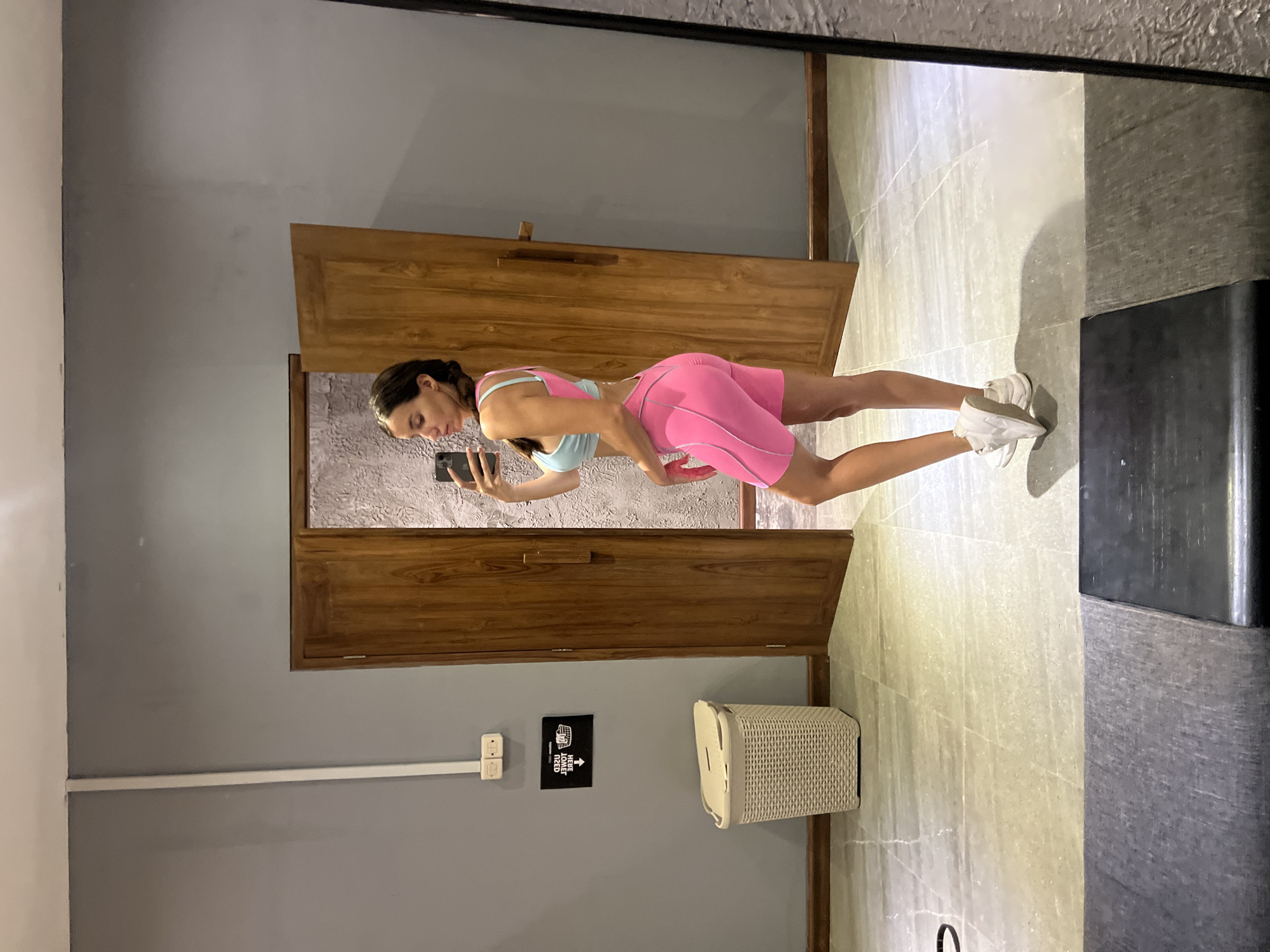 A slender woman in pink bike shorts, a light blue tank top, and white athletic shoes stands in front of a mirror, holding a cell phone, presumably taking a photograph of herself. The reflection shows a bathroom or changing area in North Kuta, Indonesia, with gray walls, wooden doors, and light tile flooring. A woven laundry basket and a sign with an arrow are visible on the wall. A black fixture is visible in the mirror's corner.