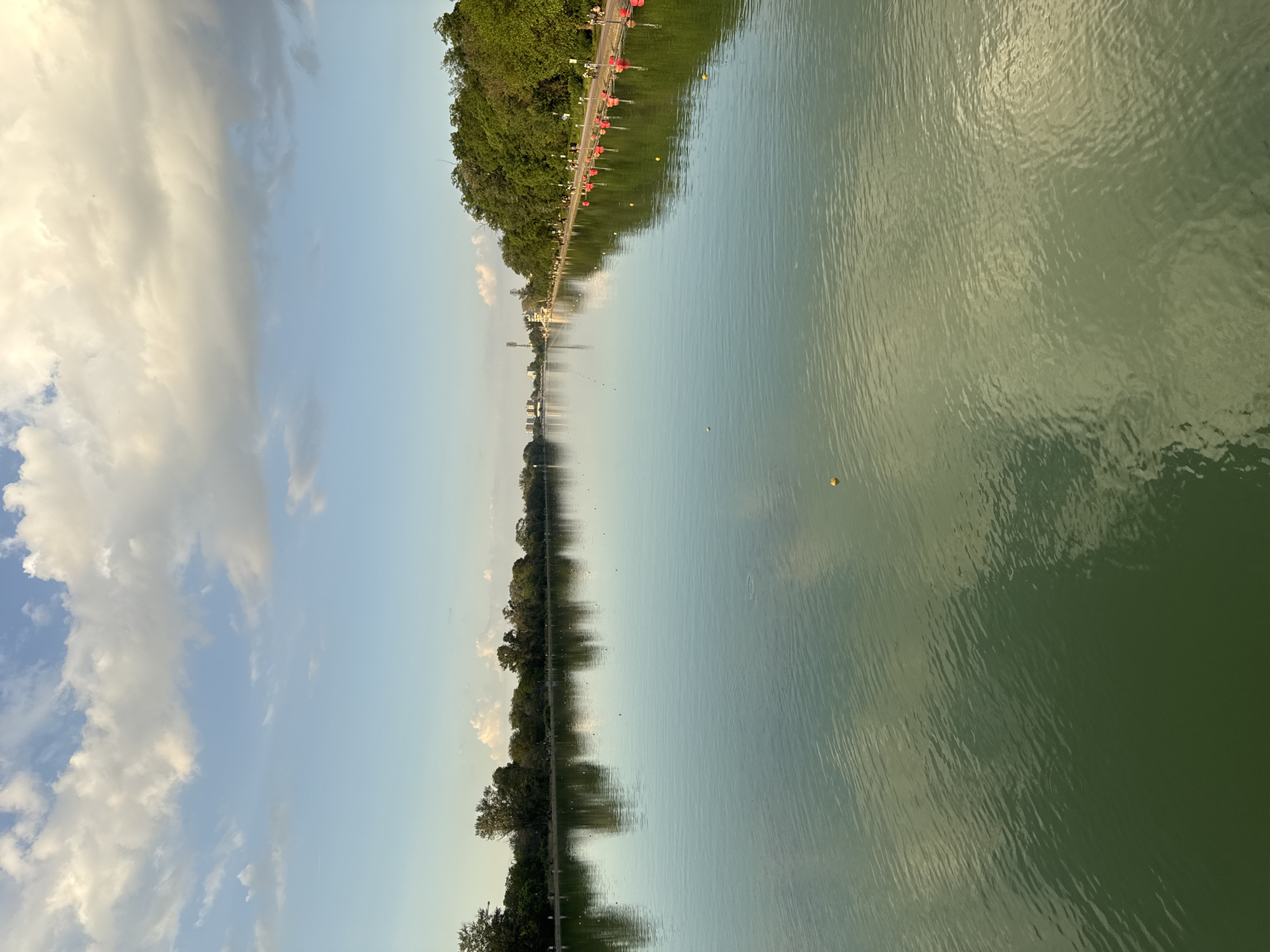 The image captures a serene landscape with a body of water, possibly a lake or river, taking up a significant portion of the frame. Its waters appear calm, reflecting the sky and the adjacent landscape.
On the left side, a vibrant blue sky is adorned with fluffy, white clouds. A line of trees is lining the bank along the left side, creating a natural boundary between the sky and the water.
The opposite bank features a road with red, marker buoys, suggesting a designated course for water sports. Beyond the road, lush greenery covers the landscape, indicating a park or natural area. In the distance, buildings and structures are visible.
Based on the landscape and the layout, the image is likely depicting the rowing canal in Plovdiv, Bulgaria. The canal is used for training and competitions.