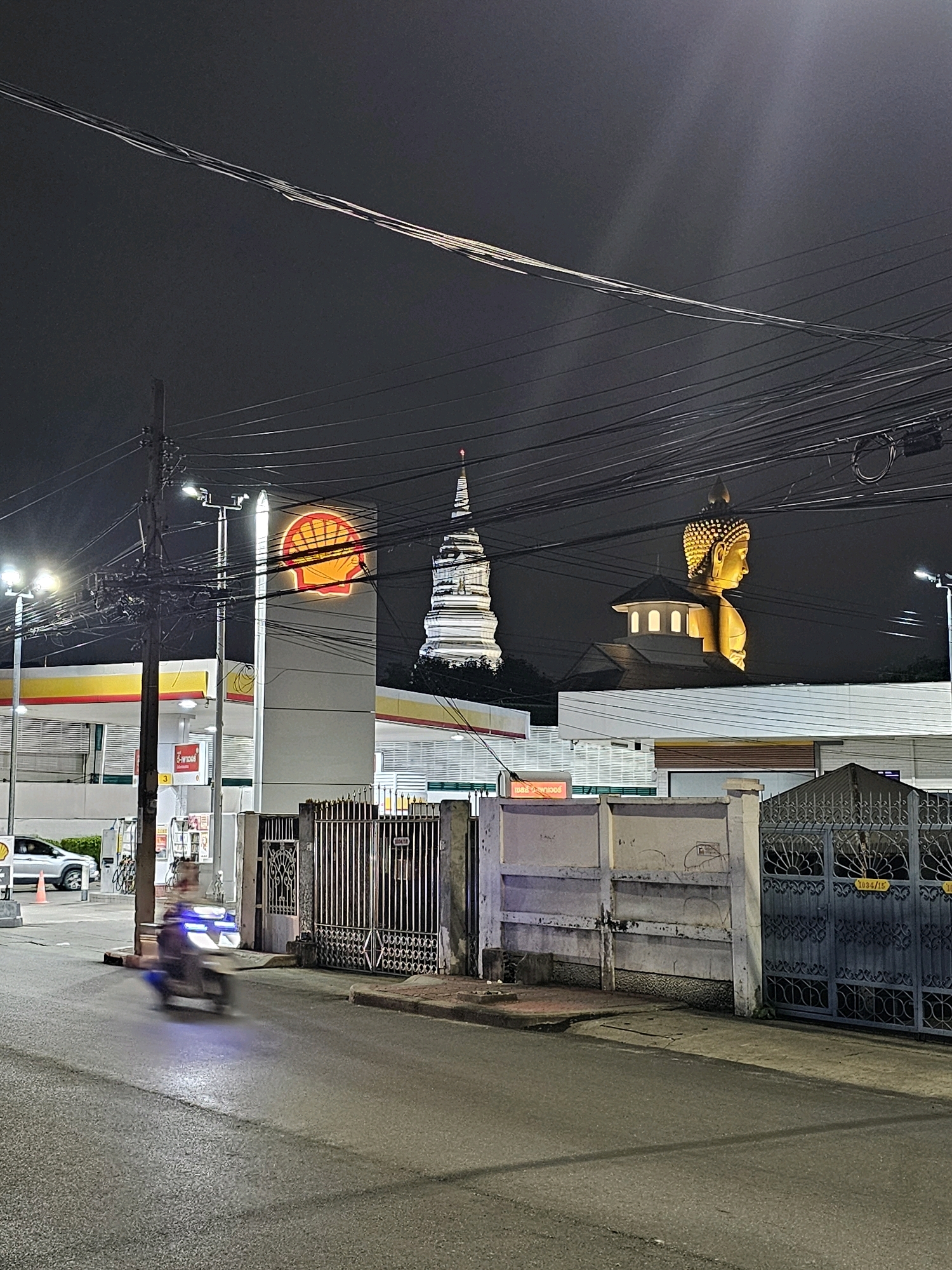 The image shows a nighttime street scene in Bangkok, Thailand. A Shell gas station is prominently featured on the left, with its iconic shell logo illuminated. To the right of the gas station, a large golden Buddha statue is visible, its head and shoulders illuminated against the dark sky, positioned behind a building. Further to the right, a white stupa, a Buddhist reliquary, is also lit up. The street in the foreground is wet, reflecting the lights from the gas station. A motorcycle rider is captured in motion on the street, blurred due to movement. In the background, numerous electrical wires crisscross the sky, characteristic of urban Thailand. A metal fence runs along the side of the street, separating the road from what appears to be a property. The overall atmosphere is that of a typical urban environment at night, with commercial and religious landmarks juxtaposed.