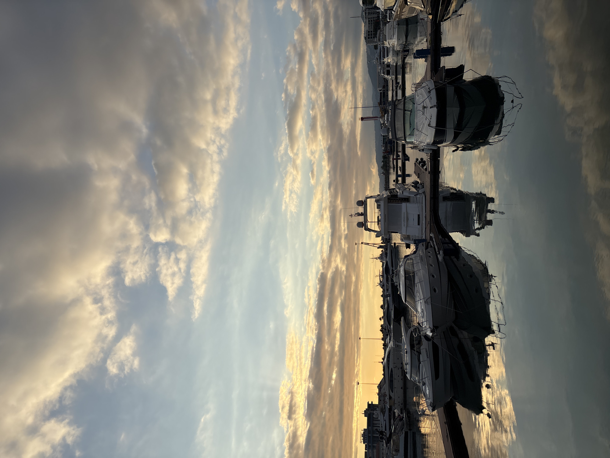 The image depicts a marina in Sochi, Russia, likely at sunset, with several yachts moored. The sky is filled with dramatic clouds, some tinged with the warm hues of the setting sun, while others are a darker gray. Rays of sunlight pierce through the clouds, creating a luminous effect. The calm water of the marina reflects the sky and the boats, adding depth to the scene. In the background, buildings and the silhouette of distant mountains are visible. The boats are varied in size and design, suggesting a mix of private and perhaps charter vessels. No people are clearly visible in the image. The overall atmosphere is serene and picturesque.