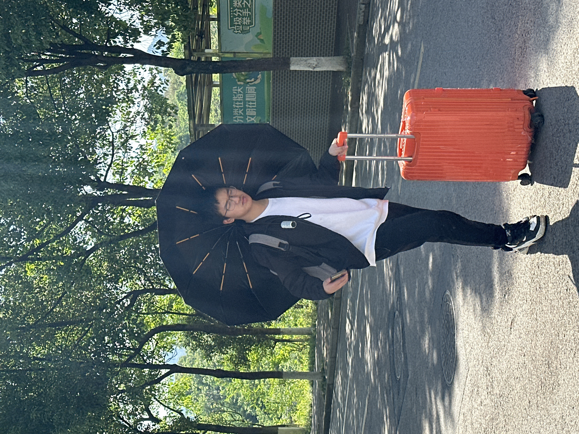 The image captures a young East Asian man walking outdoors on a sunny day in Jiangjin, China. He is pulling a bright orange hard-shell suitcase with an extended silver handle in his right hand and holding an open black umbrella in his left hand, angled for sun protection. He is dressed in a white t-shirt, a black jacket, black pants, and black and white sneakers. Glasses rest on his nose, and his eyes appear closed or half-closed, possibly indicating fatigue or focus on his smartphone, which he holds in his hand. A black backpack's straps are visible under his jacket.
The man is on a paved surface, likely a road or path, with strong sunlight casting distinct, dappled shadows from overhead trees onto the ground. Two manhole covers are visible on the pavement. To the left, a dense canopy of green trees creates a lush background. To the right, behind the man, a brick wall is visible, on which two green signs with white Chinese characters are mounted.
Visible text on the signs includes "垃圾分类" (Waste classification). Another partially visible sign reads "分类在指尖" (Sorting at your fingertips) and "文明在瞬间" (Civilization in an instant). The bright orange suitcase provides a pop of color against the otherwise subdued palette of the man's attire and the environment. The scene depicts a moment of travel or daily activity under clear skies.