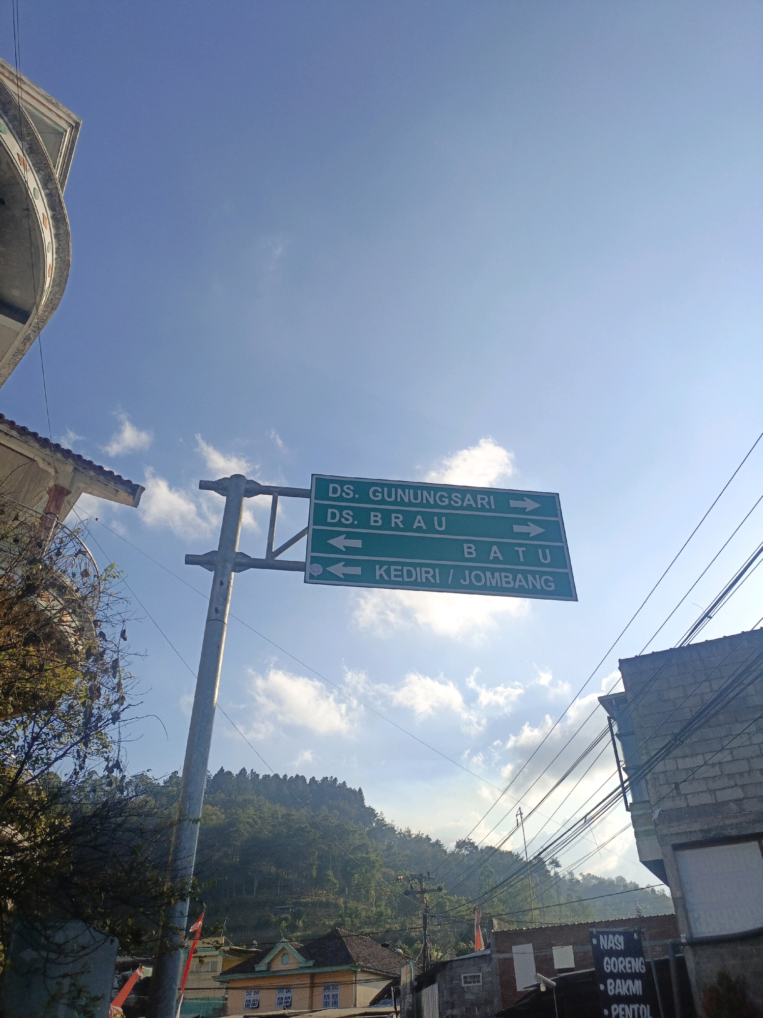 The image is an outdoor, eye-level shot of a street scene in Batu, Indonesia. The sky is a clear blue with some scattered white clouds, and the sun is bright, casting shadows. A green directional signpost is prominently displayed, indicating routes to "DS. GUNUNGSARI ->", "DS. BRAU <-", and "KEDIRI / JOMBANG <-". To the left of the signpost, a portion of a building with decorative trim is visible, along with a leafless tree and some power lines. To the right, another building constructed of brick is seen, also with power lines running across it. In the background, a densely forested hill rises under the blue sky, with several buildings visible at its base, some appearing to be residential homes with tiled roofs. The overall impression is a sunny day in a rural or semi-rural part of Batu, with signs directing travelers to various locations.