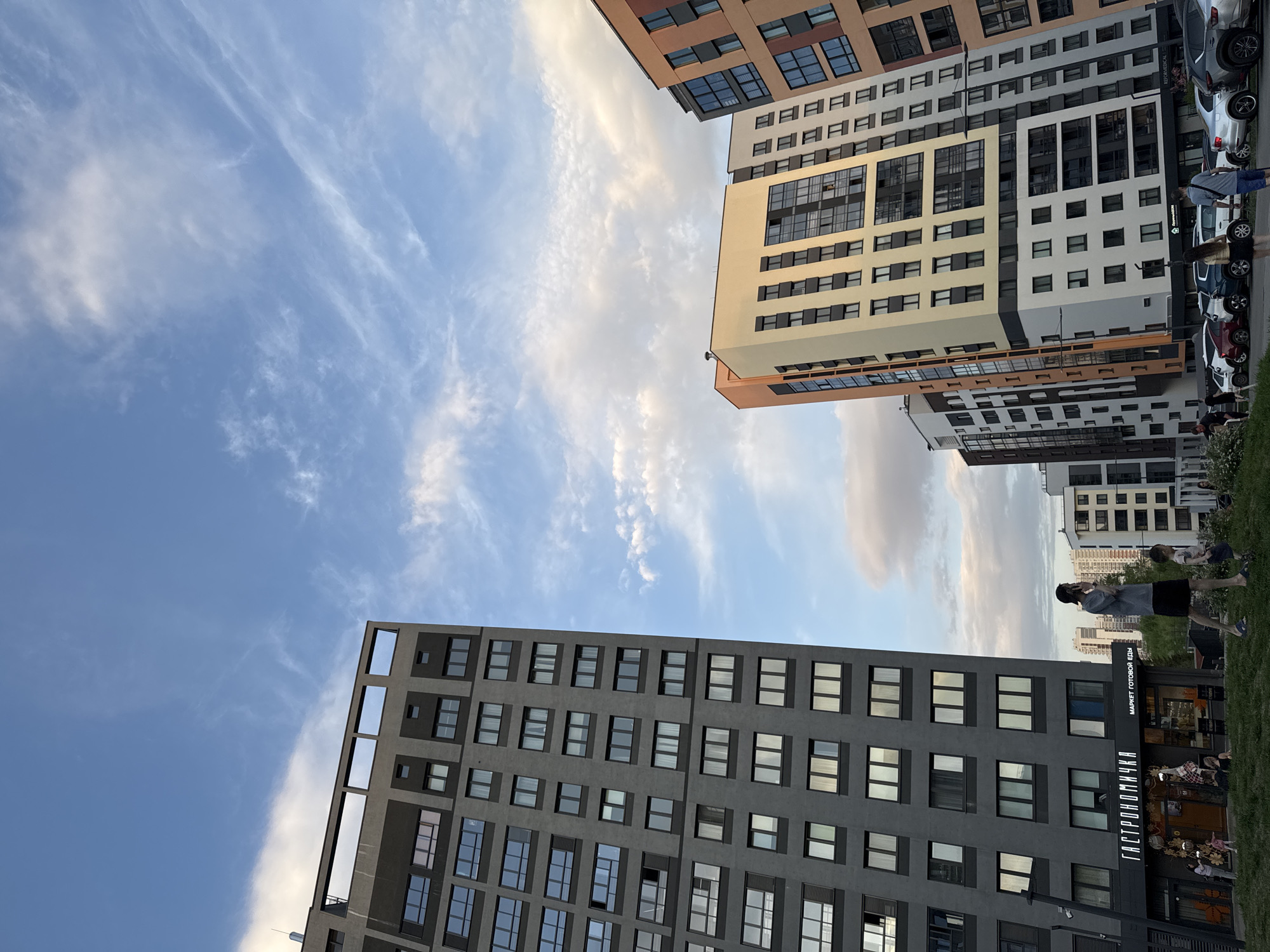 The image presents a view looking up towards several buildings against a blue sky with streaky clouds. The buildings have a modern architectural style and are multi-storied. One building is predominantly gray with evenly spaced windows. Another building features a mix of colors including tan, white, and orange. A group of people are walking beside the buildings along the sidewalk. Cars are parked along the edge of the sidewalk. There is a building with a shop called "ГАСТРОНОМИЧКА", which translated means Gastronomichka and is a store with prepared food. Based on the presence of this shop, the location is most likely in a city in Russia. Further research suggests that it is in the city of Izhevsk in Russia.