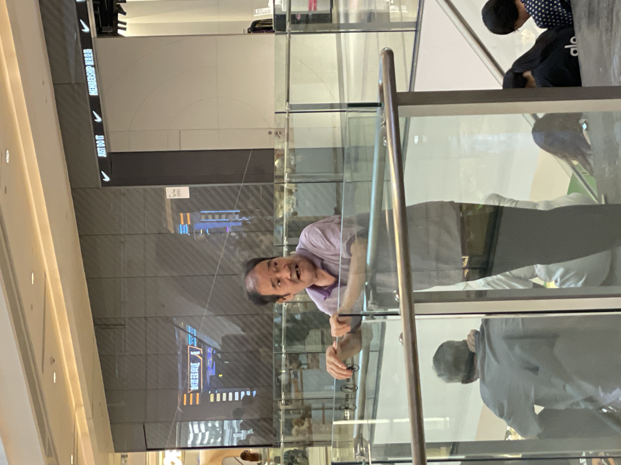 In the Yuzhong District of China, an Asian man with receding dark hair and a visible upper lip scar is standing behind a glass railing in a brightly lit, modern shopping mall. He is wearing a short-sleeved purple polo shirt and has his mouth open in a distressed expression. His left hand is resting on the railing, and his right arm is bent, with his hand near his chest. Behind him, blurred reflections show a store with neon signs and the words "JUST DO IT" and "MEMBERSHIP" with Nike swoosh logos. In front of him, several other individuals are partially visible, including a man in a grey shirt facing away from the camera, and two people, possibly women, with dark hair on an escalator in the background. The environment is clean and orderly, with a polished floor and glass partitions.