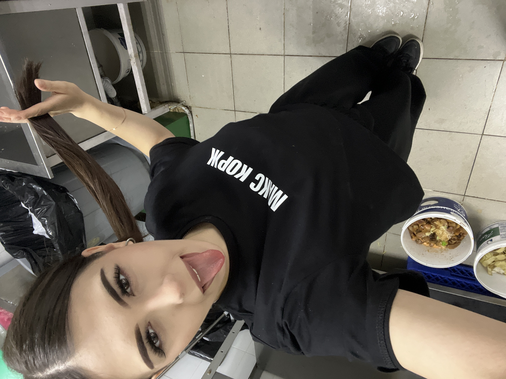 A forensic examination of the image reveals a young woman, believed to be located in Красные Баки, Russia, standing in what appears to be a commercial kitchen or utility space. She is depicted from a high-angle, selfie perspective, looking directly at the camera with her tongue extended. Her dark hair is styled in two long pigtails, one of which she holds with her right hand. She wears a black short-sleeved t-shirt with the white Cyrillic text "МАКС КОРЖ" (MAKS KORZH) emblazoned across the chest, black pants, and black sneakers with white soles. A thin, light-colored bracelet is visible on her right wrist. Her face shows defined eyebrows, dark eyeliner, and what appear to be false eyelashes.
The background consists of a light-colored tiled floor, which shows signs of moisture or spills. On the floor to her left are two white plastic buckets; one prominently features a blue label and contains what appears to be small, brown, cooked food items with some green garnish. Another partially visible bucket contains light green, possibly leafy, items. A blue plastic crate is situated between the buckets and a dark metal structure. To her right, a black garbage bag is visible near a wall with green and white panels. A metal shelf or counter runs along the right side of the frame. The overall lighting is bright and direct.
