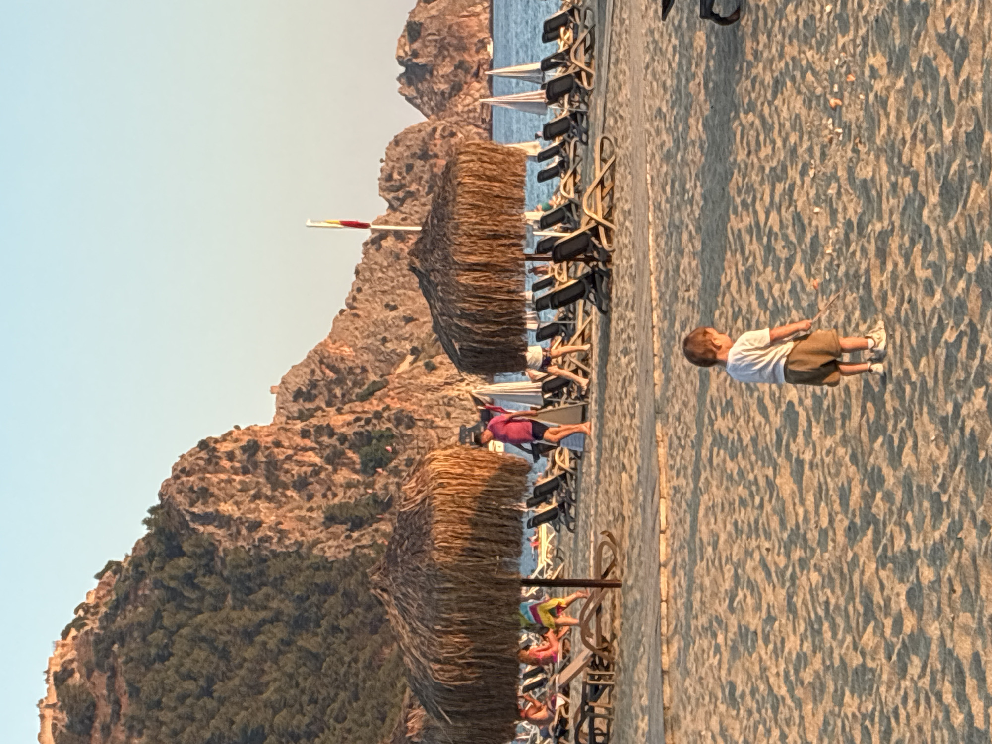This image captures a scenic beach in Alanya, Turkey, with the dramatic cliffs and ancient castle of Alanya visible in the background. The foreground is dominated by soft, golden sand, marked with gentle waves and shadows. A young child, dressed in a white t-shirt and brown shorts, stands in the middle of the sand, holding a stick and looking down at the ground, seemingly engaged in play. Behind the child, a row of empty lounge chairs and thatched umbrellas line the beach, suggesting a quiet moment before or after a busy period. The calm blue sea stretches out to the horizon, with a few white sailboats dotting the water. A flag pole with a flag is visible on the right side of the image. The warm glow of the setting sun bathes the scene in a soft, orange light, enhancing the serene and picturesque atmosphere of the Mediterranean coast.