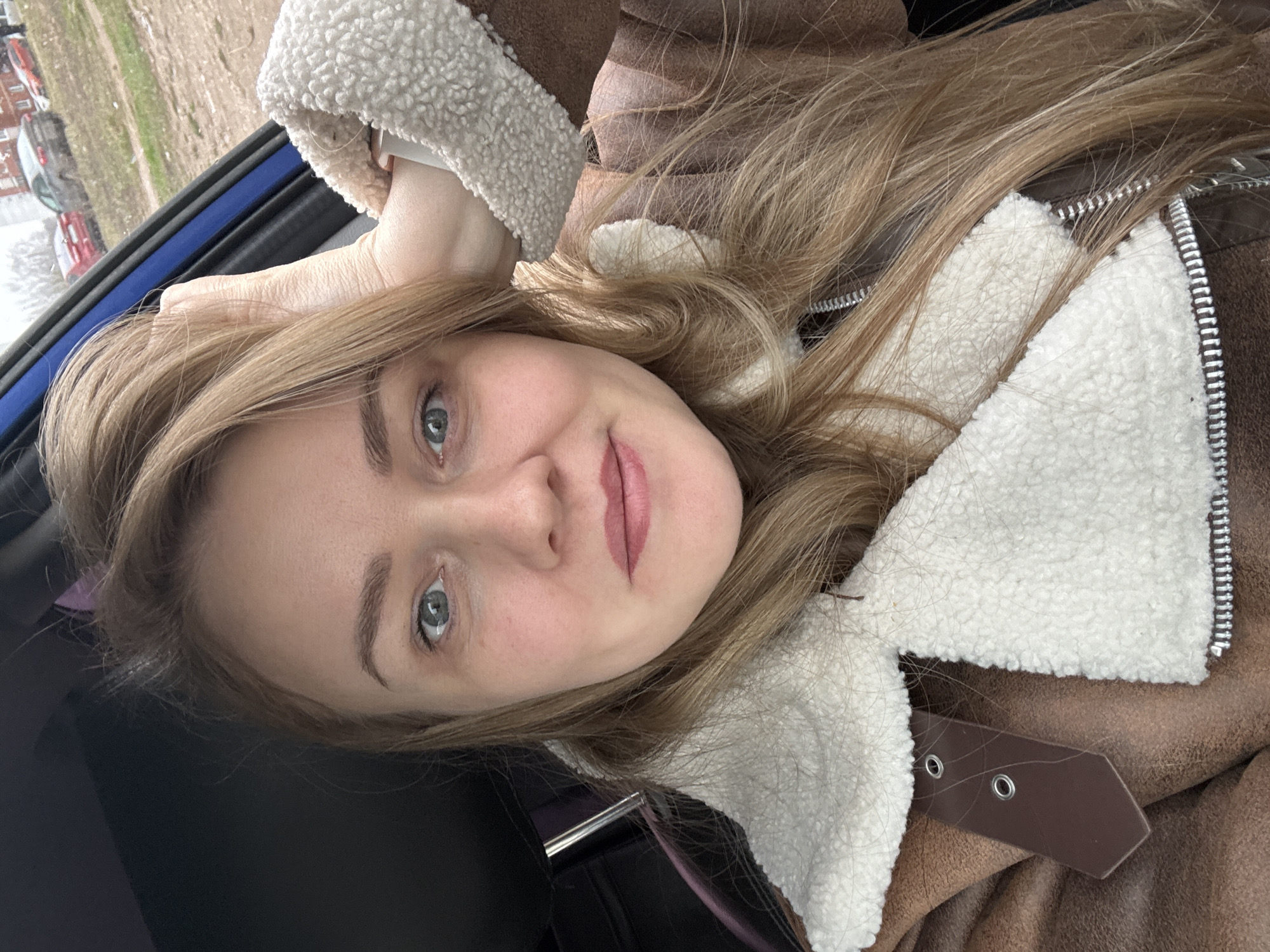 A young woman with light blonde hair and fair skin is taking a selfie. She has blue eyes and is wearing lipstick. She is wearing a brown jacket with white sherpa lining. The photo appears to be taken inside a vehicle, as evidenced by the window frame and dark interior visible in the background. Through the window, there is a view of what appears to be a cityscape, potentially suggesting the location of Chaikovsky, Russia. There are buildings and vehicles visible outside. The woman has one hand raised near her head.