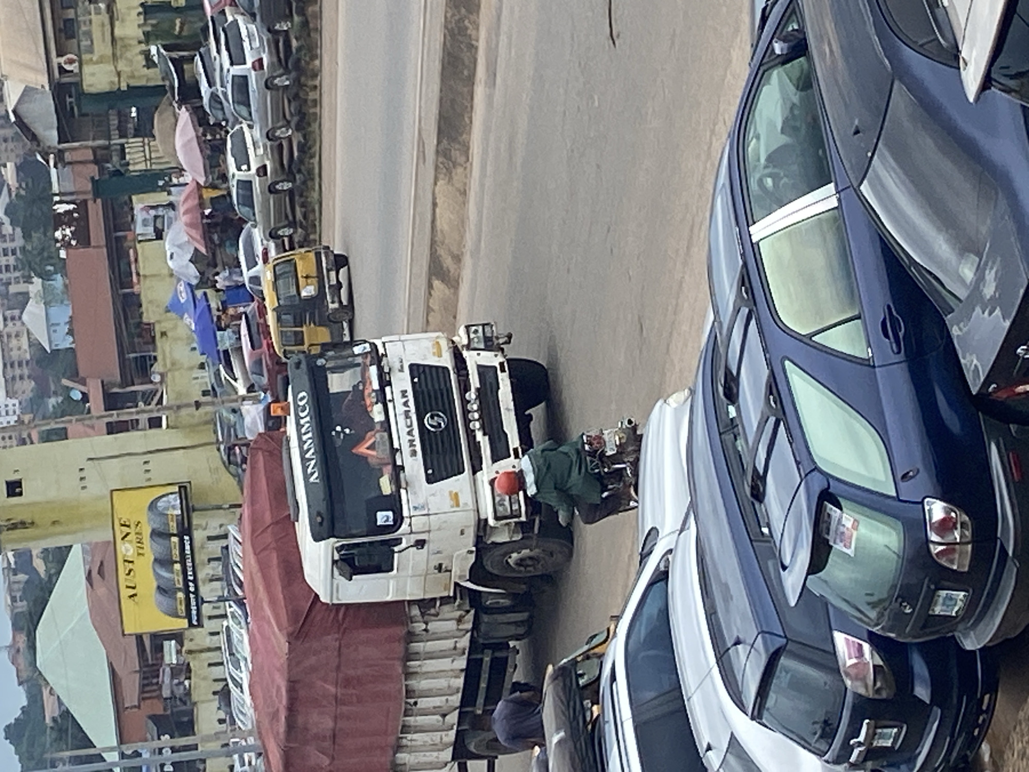 This image depicts a busy street scene in Ogbunike, Nigeria. Several vehicles are present, including a large white truck with "ANAMMCO" written on its side, a yellow auto-rickshaw, and several cars, including a prominent dark blue sedan. People are visible, with one person in a green jacket and red helmet working on the back of the white truck, and another person in the background near the auto-rickshaw. The setting appears to be a commercial area, with buildings and a billboard advertising "AU STONE TIRES" visible. The overall atmosphere suggests a typical day with traffic and commercial activity. The lighting indicates it is daytime, and the weather appears to be overcast.
