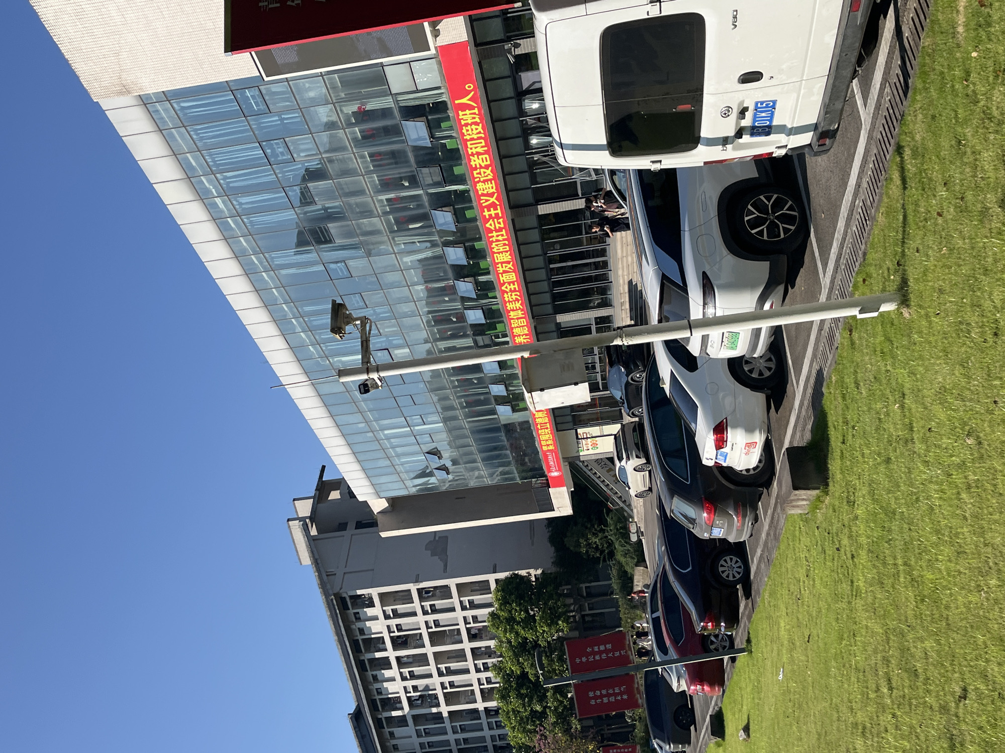 A bright, sunny daytime scene captured at an angle shows a modern, multi-story building with a glass facade and a long red banner displaying Chinese text across its lower section. To the left, a taller, older building with many windows is visible. In the foreground, a row of parked cars, including a white van with a license plate reading "B0IK15," lines a parking lot adjacent to a lush green lawn. A security camera is mounted on a pole in the center of the frame. The image appears to be taken in Jiangjin, China, likely on a campus or public area, given the style of buildings and the presence of banners.