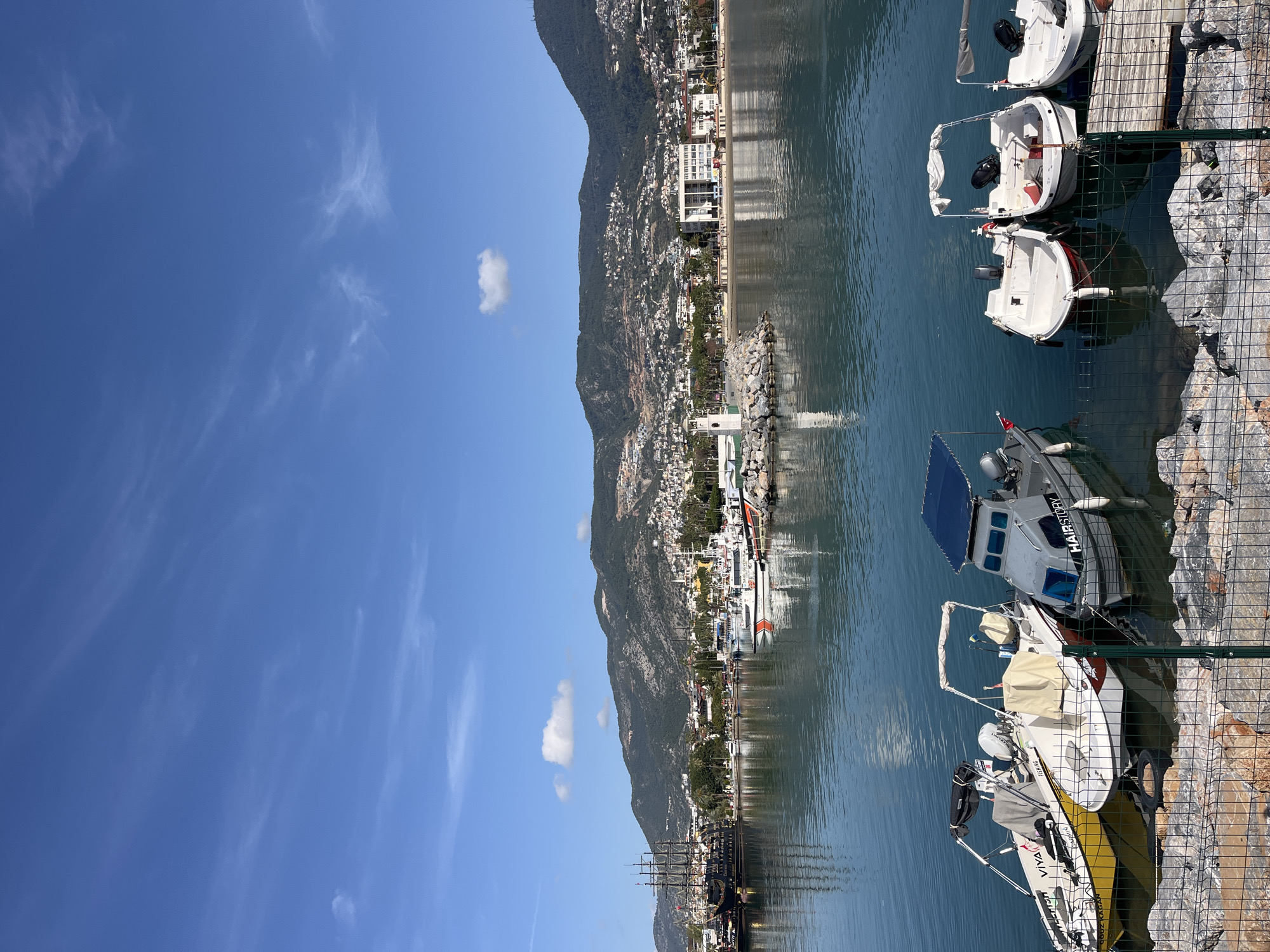 The image depicts a waterfront scene, likely the harbor in Alanya, Turkey, based on the architecture and landscape. A mountain slopes down to meet a coastline densely populated with buildings. The water surface reflects the buildings and sky. In the foreground, several small white boats are docked alongside a fenced pier. The sky is bright blue with wispy clouds. No people are visible.