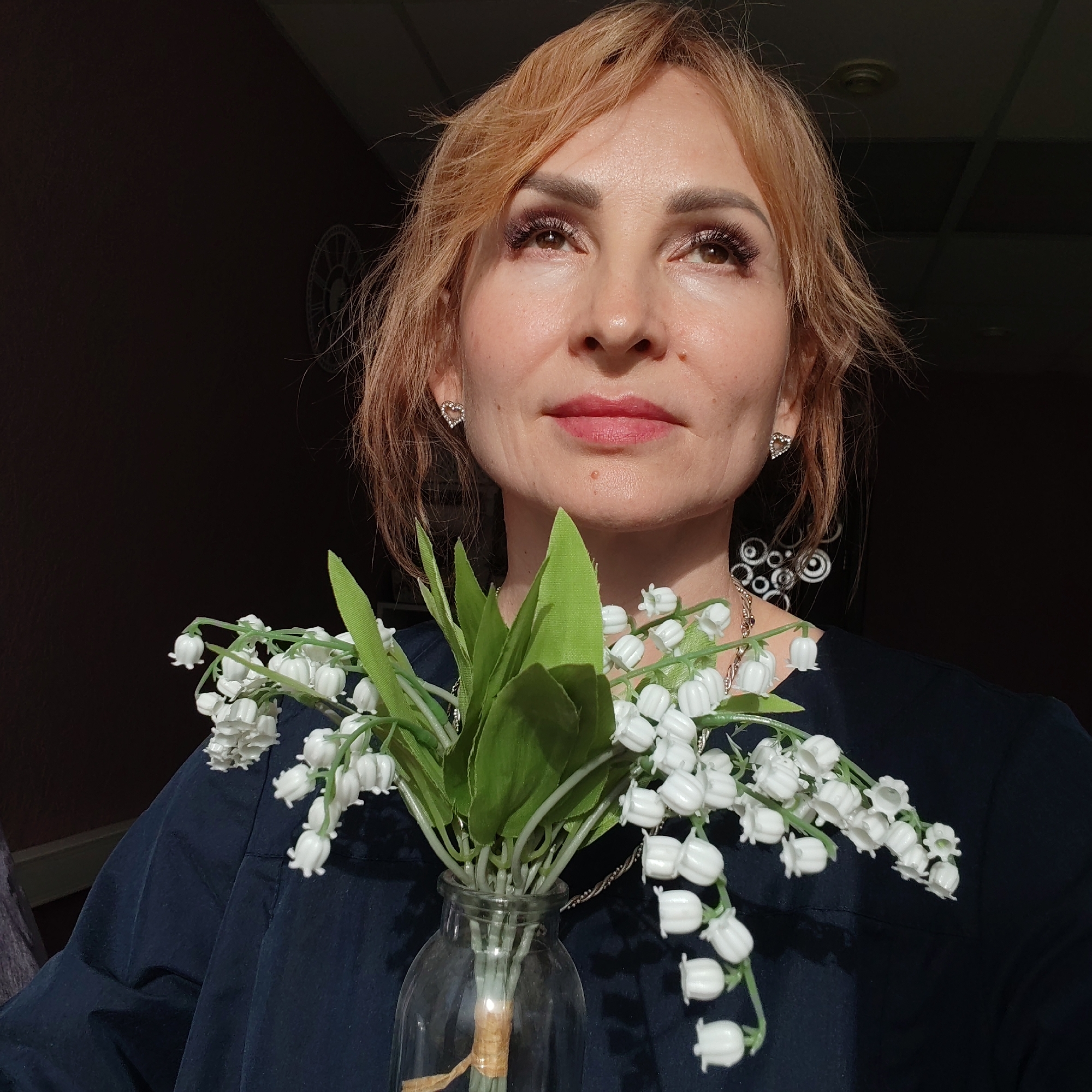 A woman with light reddish-blonde hair holds a glass vase filled with lily of the valley flowers in front of her. The woman has fair skin, is wearing heart-shaped earrings, and has light pink lipstick. She is looking upwards. Behind her is a dark background, and to the left of the background, there is a clock. She is wearing a dark blue shirt or dress. The location is Chaikovsky, Russia.