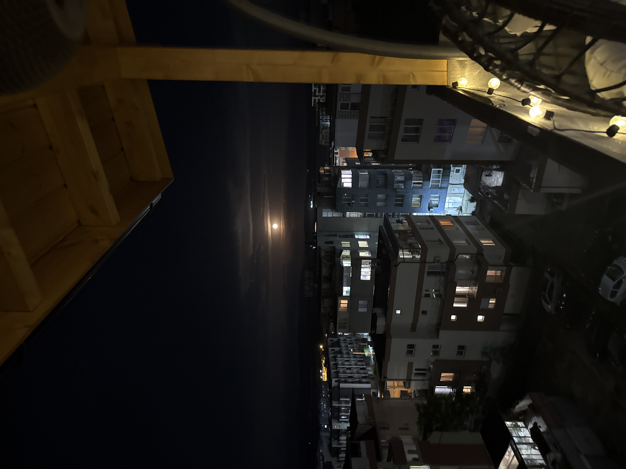Here's a detailed description of the image:
**Scene:** A night view from an elevated position, likely a balcony or rooftop. The scene depicts a cityscape at night, with a visible moon over a body of water (likely the Black Sea).
**Location:** Based on the architecture and context of the image, the location appears to be Burgas, Bulgaria.
**Objects:**
* **Buildings:** Multiple apartment buildings with illuminated windows, indicating residential activity.
* **Street Lights:** There is a street light on the coast
* **Cars:** Several cars are parked in the streets below.
* **Moon:** A bright, round moon is visible over the water.
* **Balcony Elements:** Parts of the balcony structure are visible in the foreground, including wooden beams and string lights.
* **Water:** A wide expanse of dark water stretches toward the horizon.
**Environment:**
* **Lighting:** Predominantly dark, illuminated by moonlight and artificial lights from the buildings and streets.
* **Atmosphere:** Calm night ambiance.
* **Weather:** The sky is partially cloudy, with a patch for the Moon.