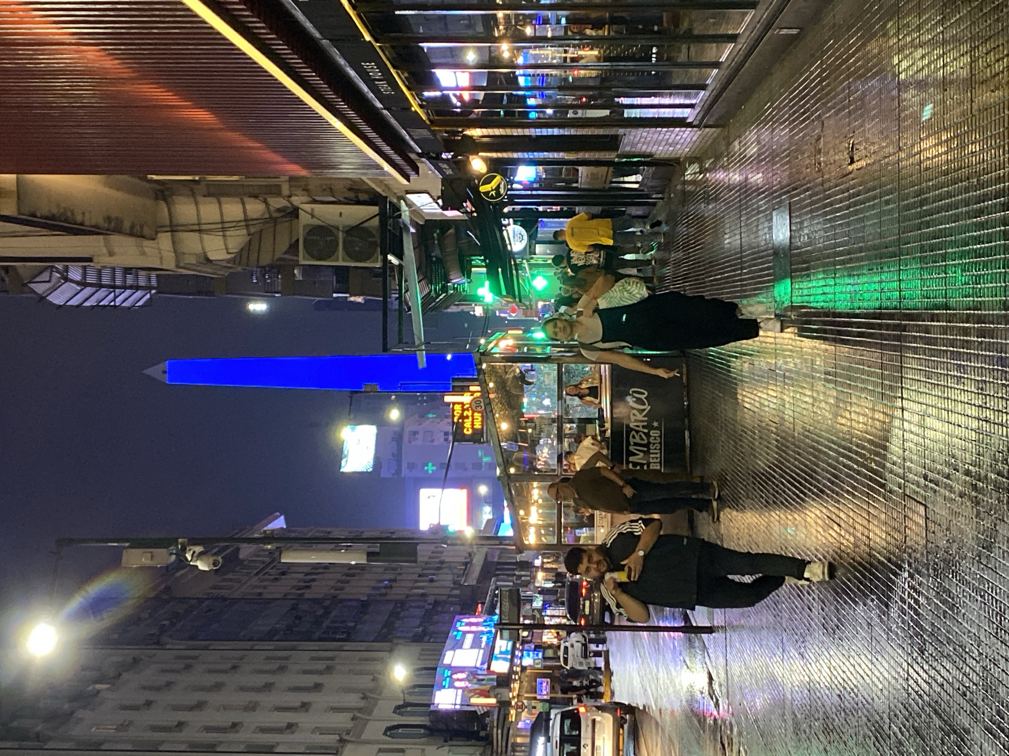 This nighttime image depicts an urban street scene in Buenos Aires, Argentina, with the Obelisco prominently illuminated in blue in the background, indicating proximity to Avenida 9 de Julio and Avenida Corrientes. The street and sidewalks are wet and highly reflective, suggesting recent rain.
In the foreground, two individuals walk on a textured metal grate sidewalk. On the left, a male subject, dressed in a dark t-shirt with white stripes on the sleeves and dark pants, holds a yellow beverage container to his mouth. On the right, a female subject, wearing a dark jumpsuit and light-colored shoes, carries a light bag over her left shoulder and holds a lit cigarette in her right hand. She looks slightly to her right.
To the right of the female subject, a commercial establishment with large glass windows and a "STEAK HOUSE" sign above is visible. Further down, an outdoor seating area enclosed by glass panels bears a partially visible sign "EMBARCO BELISCO," where several people are gathered, including a man in a dark green shirt. Multiple other pedestrians are visible further along the street.
The street itself is wet and reflects various lights from the surrounding environment, with several vehicles, including a white van, visible. Tall buildings line both sides of the street. The building on the left is lighter-colored with multiple windows. The building on the right features a reddish, vertically slatted facade with visible air conditioning units mounted externally. A lamppost on the left side of the street holds a security camera. Numerous vibrant neon signs, streetlights, and building lights illuminate the scene, casting colorful reflections across the wet surfaces. Green illuminated cross signs are visible further down the street.