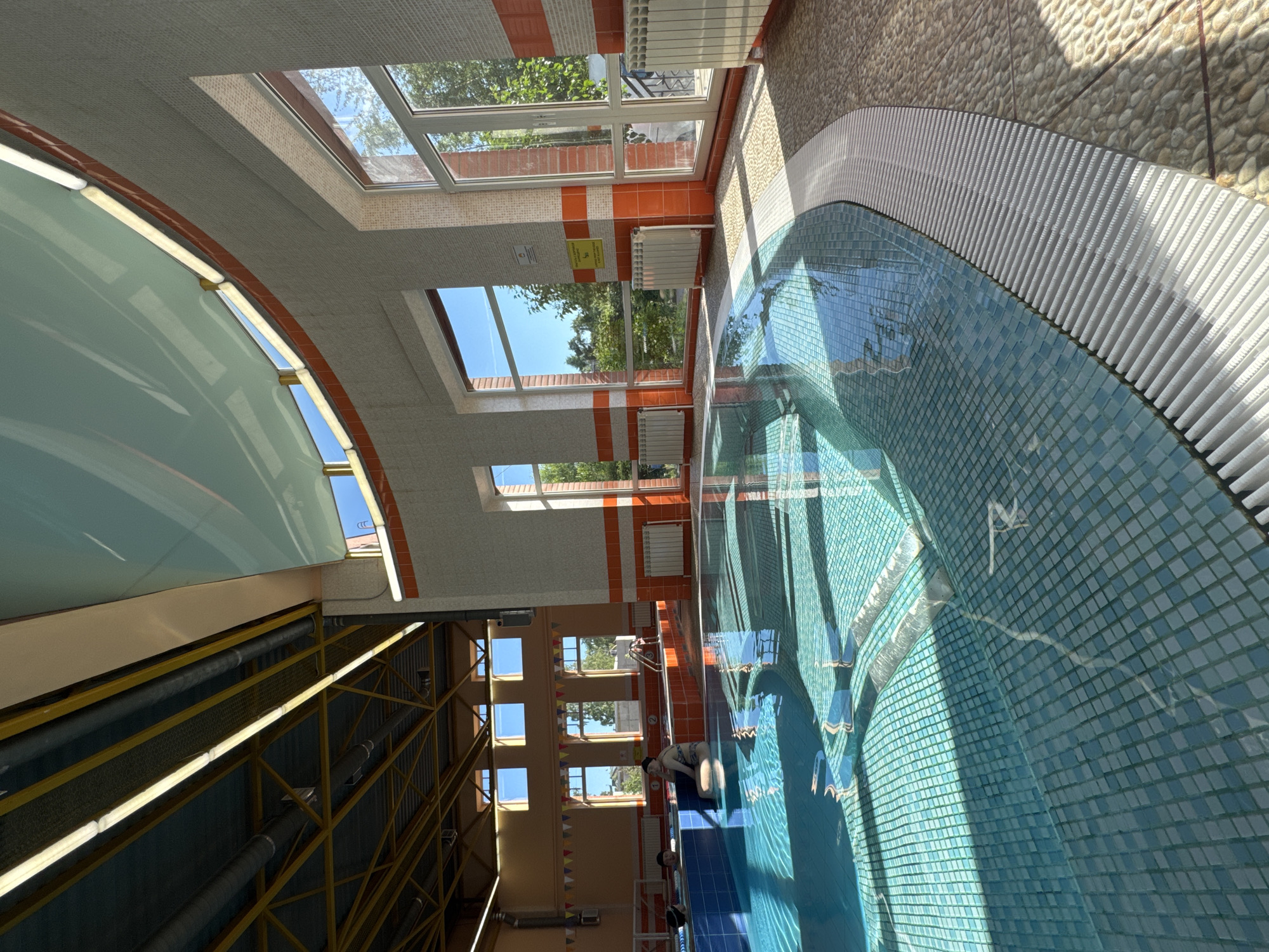 The image depicts the interior of a public swimming pool facility. The pool itself is kidney-shaped and tiled with small, light blue square tiles. Sunlight streams in through large windows, casting geometric shadows on the water's surface. The pool area is surrounded by a floor made of small, rounded pebbles and a white, ribbed border.
To the left, a raised section of the pool, also tiled in blue, contains a few individuals. One person, wearing a patterned swimsuit, sits with their back to the camera in this raised section. Another person is partially submerged in the main pool area.
The walls of the facility are a neutral color, adorned with horizontal bands of orange tiles. Large windows offer views of greenery and blue sky. Radiators are visible beneath the windows. The ceiling features exposed beams and lighting fixtures, suggesting an industrial design. String lights with small flags are visible in the background on the left side of the image. The overall impression is of a well-lit, modern indoor swimming pool.
This facility is located in Saint Petersburg, Russia.