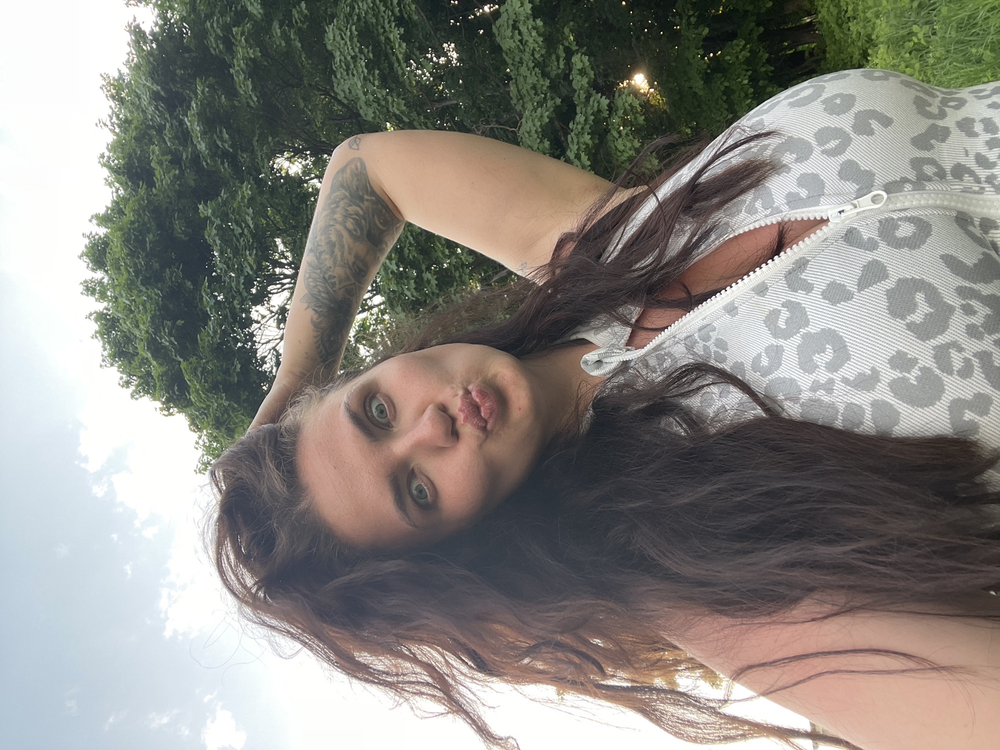 A woman with long brown hair, light eyes, and a tattoo on her left arm poses for a selfie in an outdoor setting. She is wearing a patterned zip-up top and making a kissing face. The background includes trees, suggesting a park or natural area. The location cannot be determined as Ostrov, Russia, as there are no indications of the location in the image.