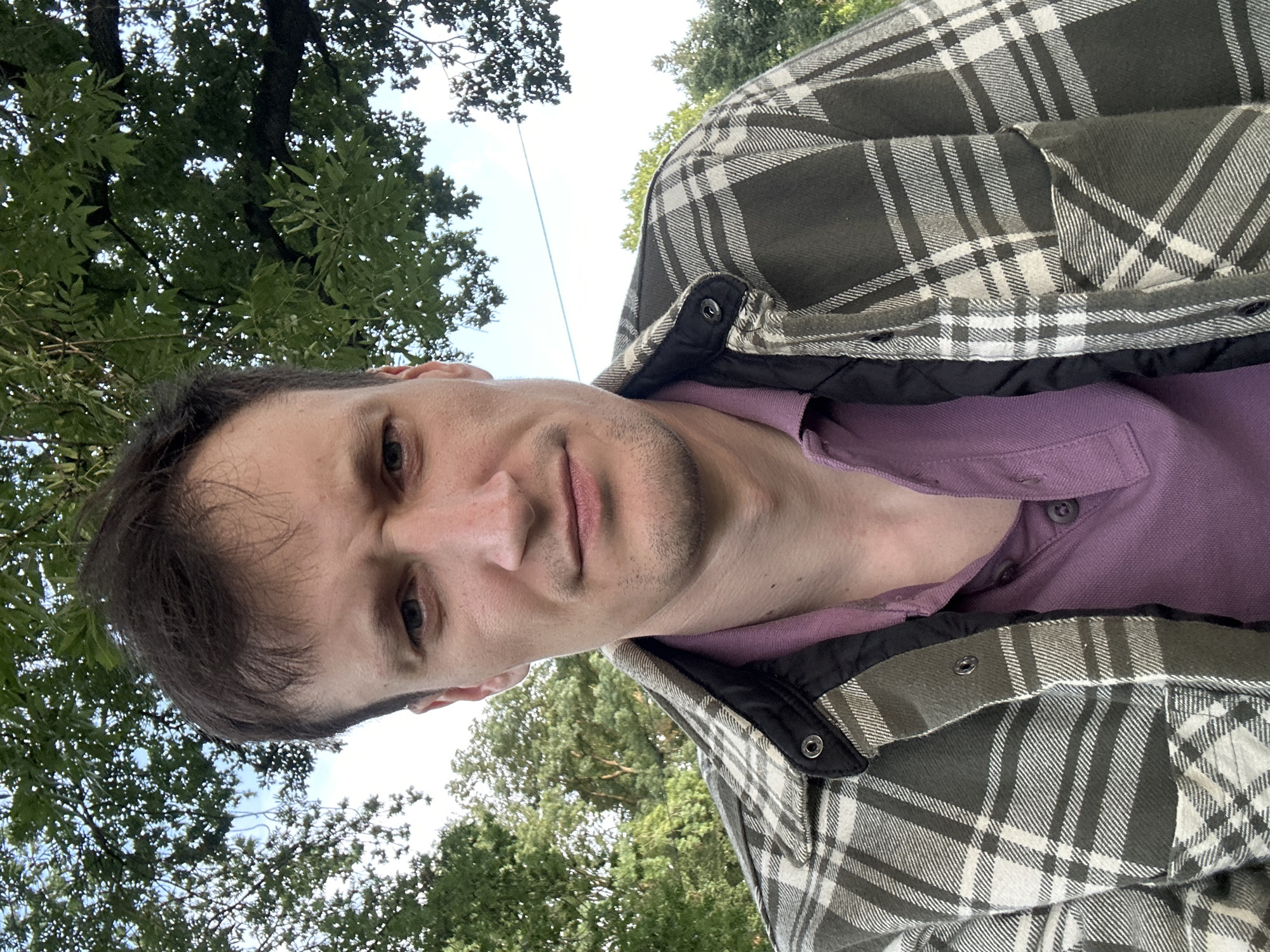 A medium shot shows a man with dark hair and blue eyes looking directly at the camera. He is wearing a purple polo shirt and a green and white plaid jacket. The background is out of focus, showing green trees and a blue sky. The image appears to be taken outdoors, possibly in a park or rural area. The man's facial expression is neutral, with a slight furrow in his brow. The lighting suggests it is daytime. There are no other people or animals visible in the frame. The man is positioned slightly off-center to the left, with the majority of the frame occupied by his upper body and head.