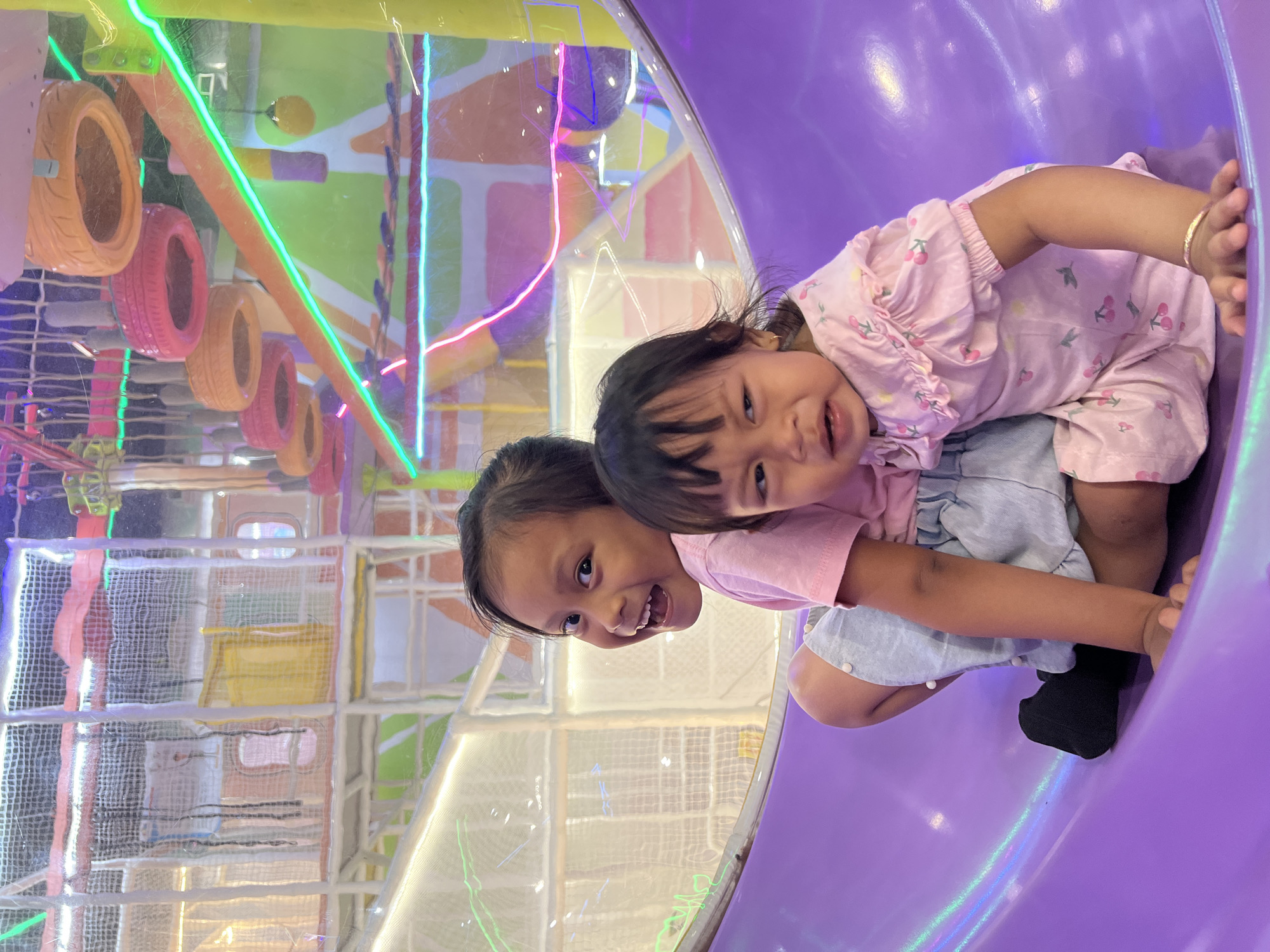 Two young children, a girl and a toddler, are sitting on a purple slide inside an indoor playground. The girl, on the left, is smiling broadly and looking towards the camera, wearing a pink t-shirt and light blue shorts. The toddler, on the right, is crying and looking away, dressed in a pink shirt with a cherry pattern and a matching pink skirt. The background of the playground is brightly colored with various structures, netting, and neon lights, suggesting a vibrant and playful environment. The location is specified as Kecamatan Pati, Indonesia, indicating an indoor entertainment facility within that region.