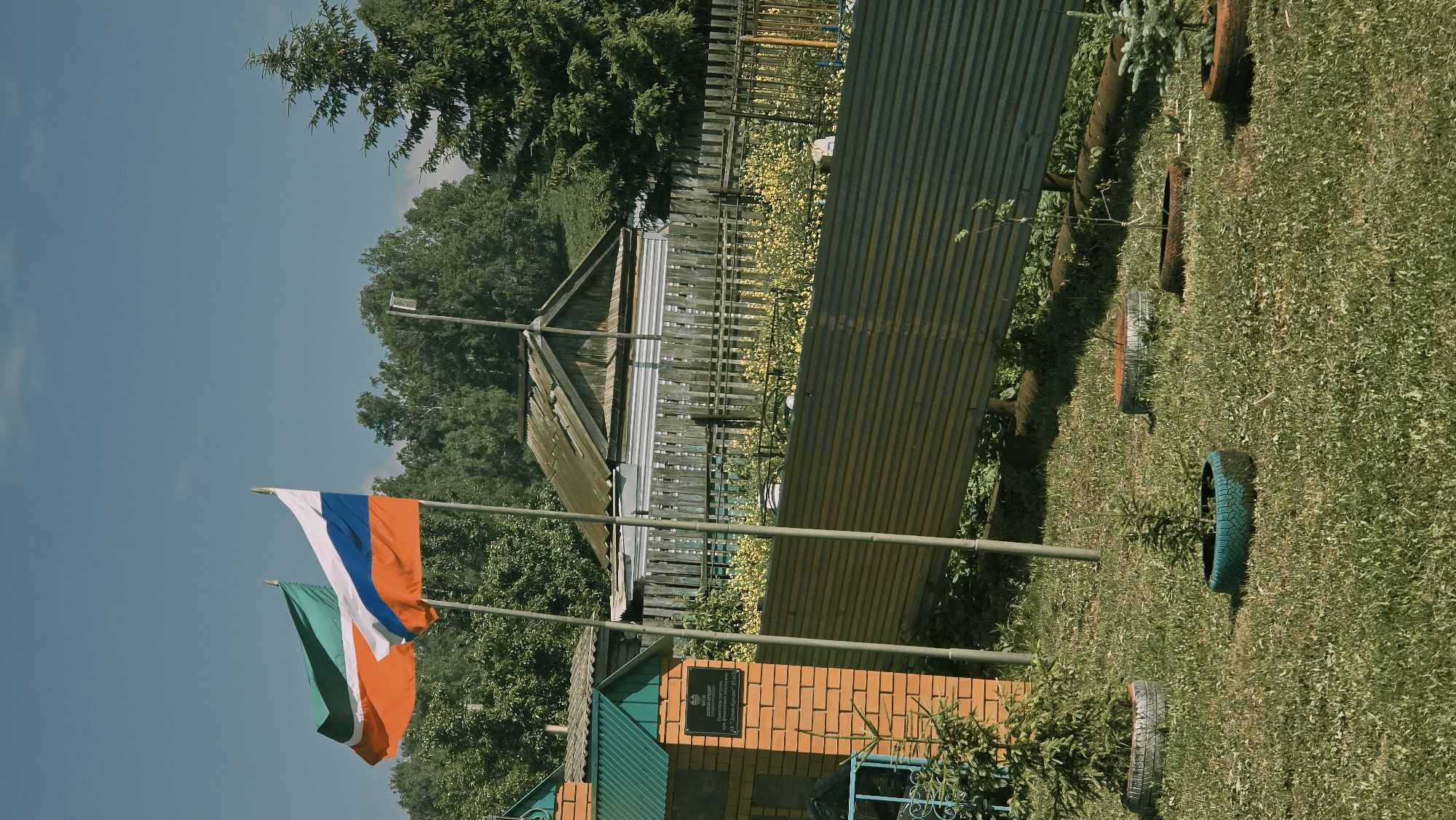 The image depicts an outdoor scene under a clear blue sky, with green trees forming a backdrop. In the foreground, two flags are prominently displayed on poles. The flag on the left is green, white, and orange, while the flag on the right is the Russian tricolor, featuring white, blue, and red horizontal bands. To the left of the flags, a brick structure with a green roof is visible, featuring a plaque with text. In front of this structure, a young evergreen sapling is planted in a tire. Several other tire planters with saplings or small trees are arranged on the grassy lawn. A corrugated metal fence runs horizontally across the middle ground, behind which a wooden fence and a simple wooden house with a weathered roof are visible. The overall environment suggests a rural or semi-rural setting.