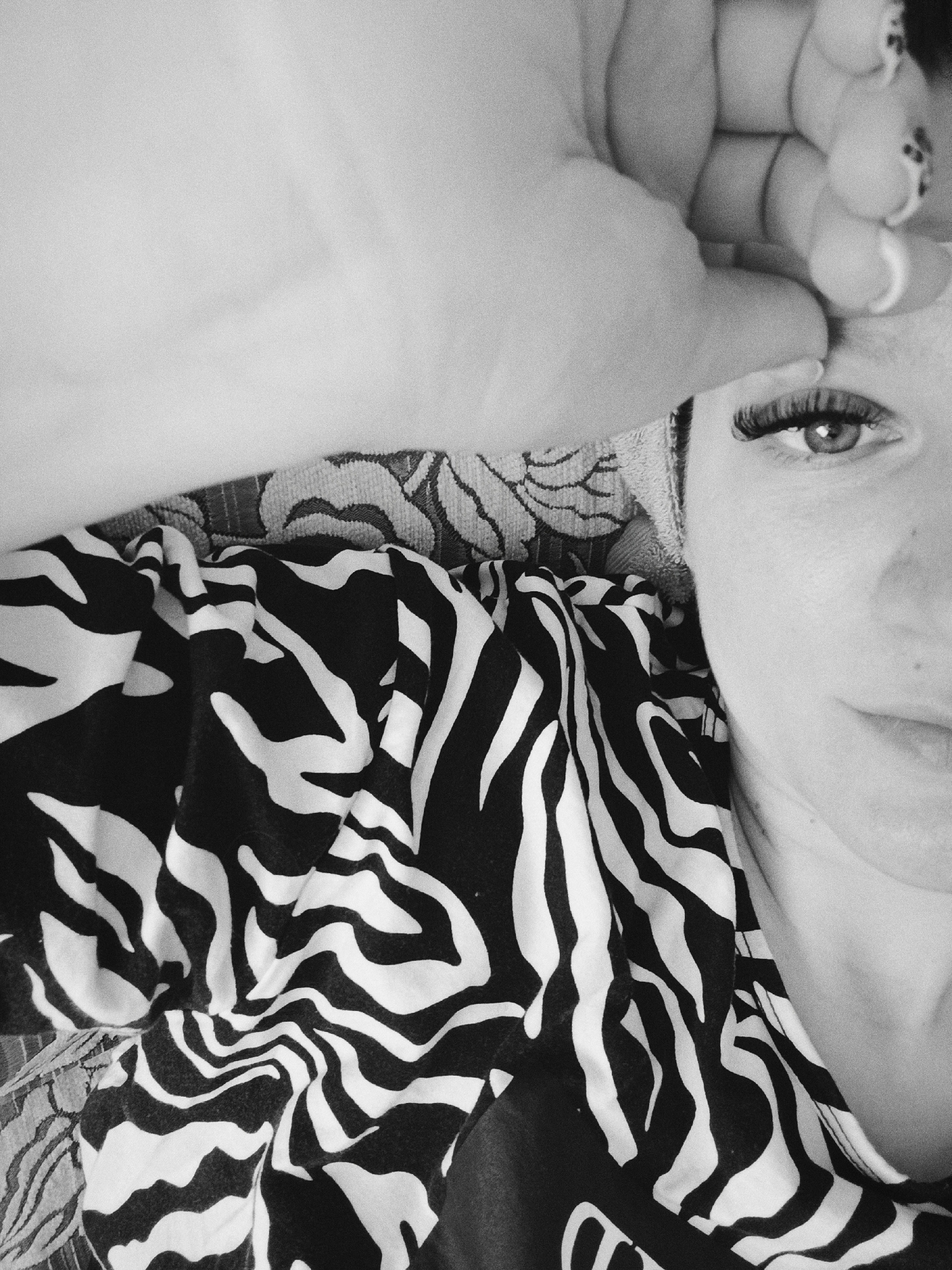 A black and white close-up photo shows a woman lying down, with her face partially visible. Her right eye is open, looking directly at the camera. She has dark eyelashes. Part of her right hand is on her forehead. She wears a shirt or blouse with a zebra print pattern. The background suggests a patterned fabric surface like a bed or couch. There is no information to confirm it is in the city of Tobolsk, Russia.