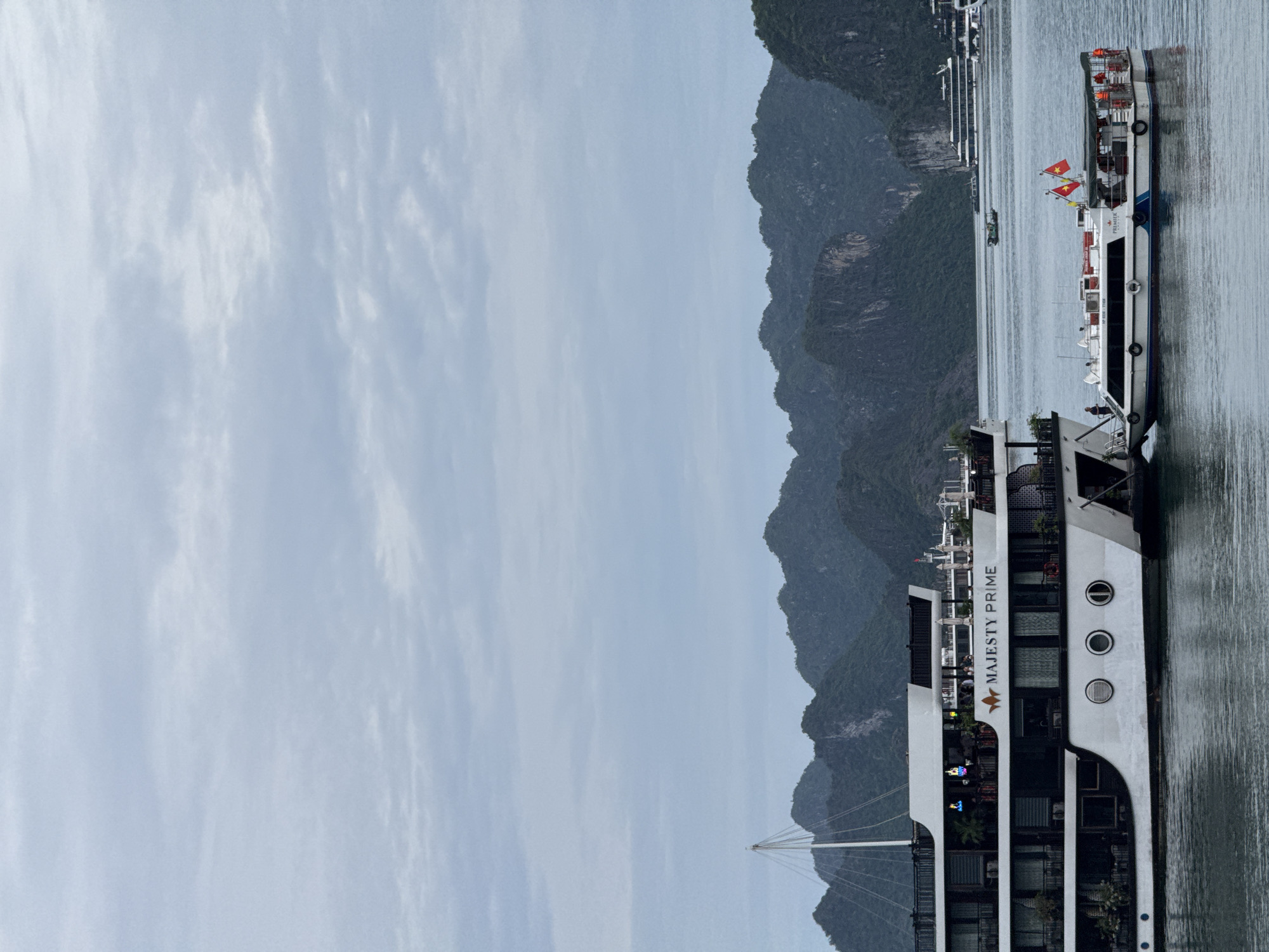 The image shows a side view of the "MAJESTY PRIME" cruise ship docked in a body of water, with a mountainous landscape in the background. The sky is overcast, indicating a cloudy day. The ship is white with dark windows and several decks, featuring outdoor seating areas. Two Vietnamese flags are visible on the top deck. Another smaller boat is also present in the water. The scene appears to be a serene, possibly tourist-oriented location, given the presence of a cruise ship and the dramatic natural scenery. The water is calm, reflecting the muted light of the day. The overall impression is one of a peaceful setting in a coastal or island environment.